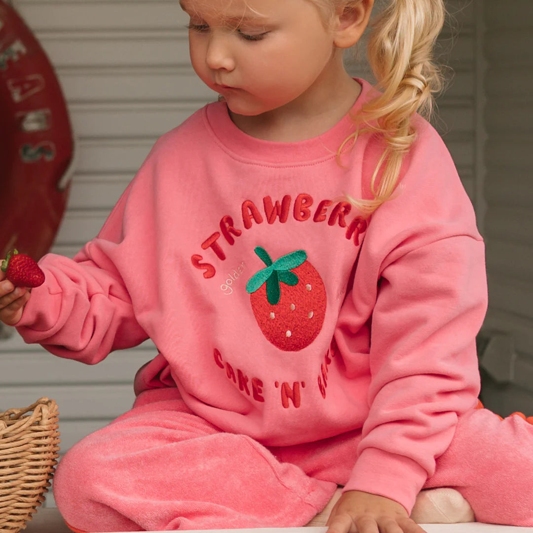 A young child in the GOLDEN CHILDREN Golden Children - Strawberry Cake Sweater Meringue Pink, made of cozy french terry fleece cotton with a strawberry design and relaxed fit, sits holding a strawberry with a basket nearby.