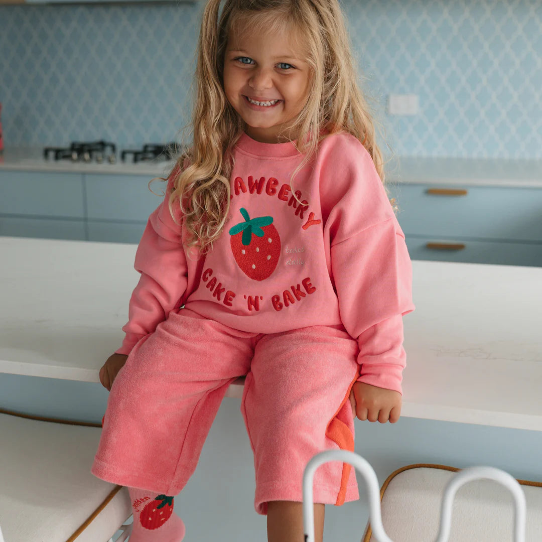 A young girl with long blonde hair, wearing the GOLDEN CHILDREN Golden Children - Strawberry Cake Sweater Meringue Pink, sits smiling on a white kitchen counter with light blue cabinets in the background.