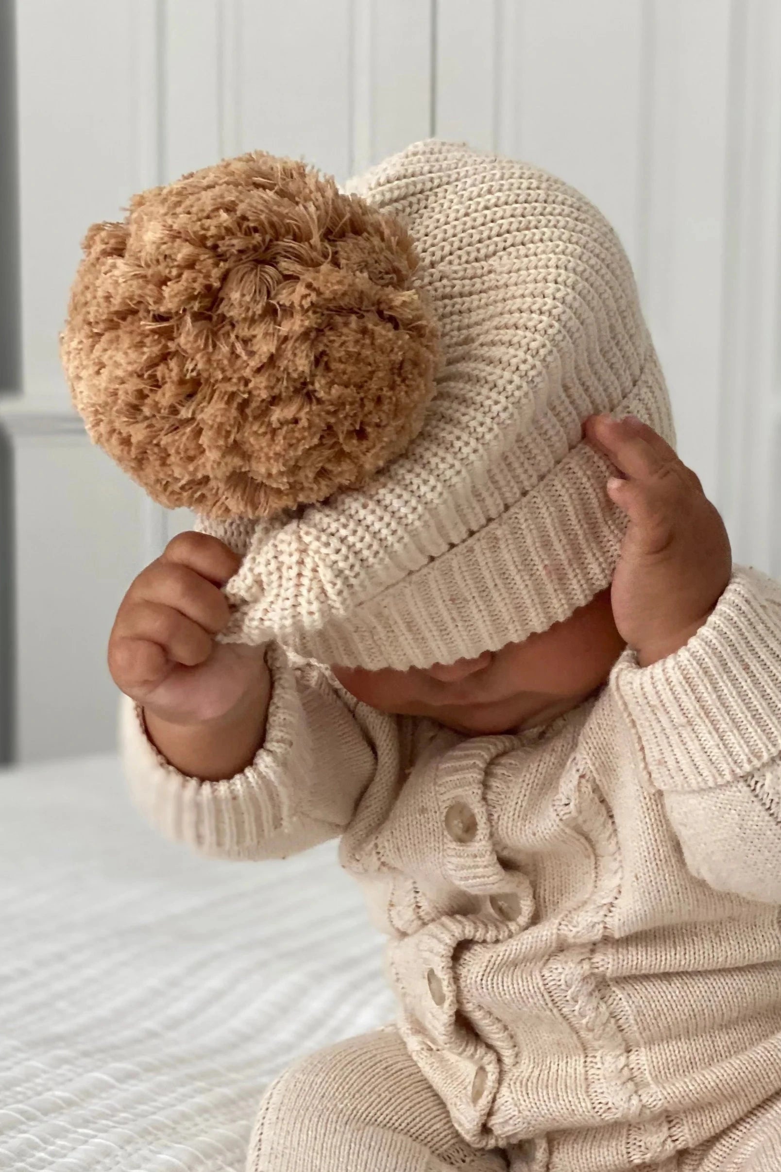 A baby in a ZIGGY LOU Beanie Biscotti Fleck and a matching sweater is sitting indoors. The baby is pulling the beanie down over their eyes with both hands, partially concealing their face. The softly lit background adds an out-of-focus touch to the scene.