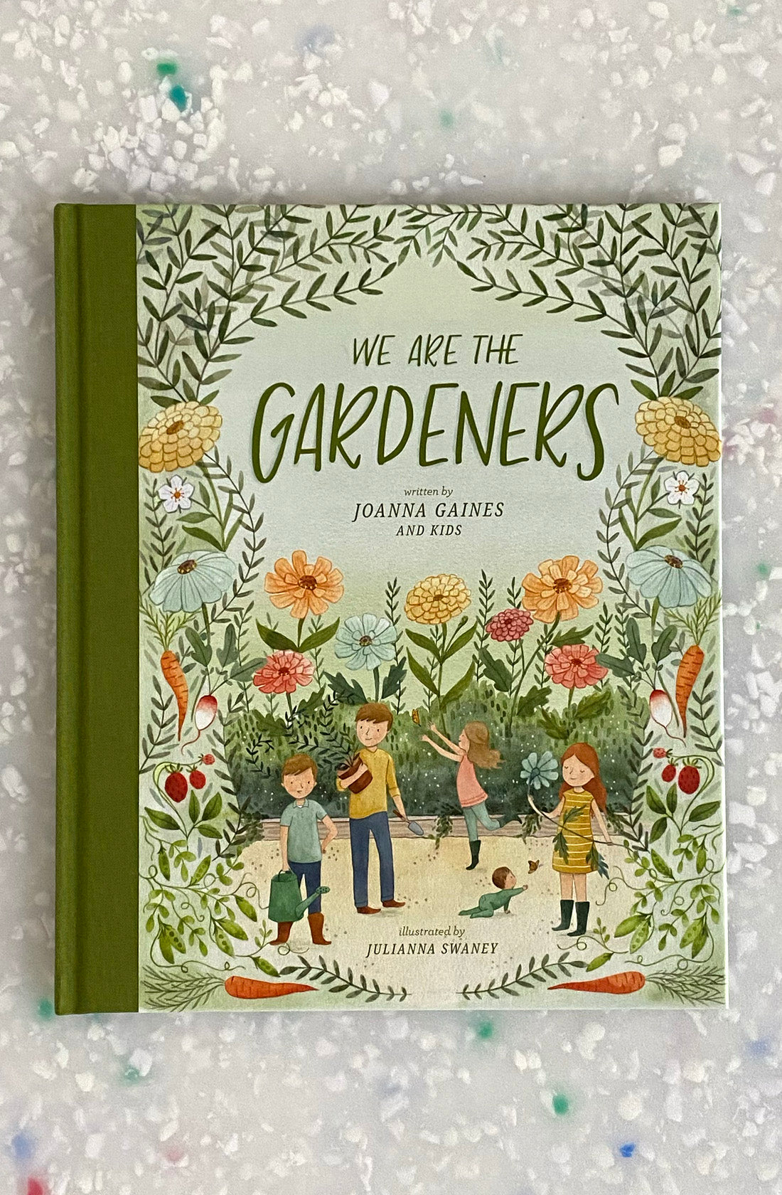 The image showcases the cover of "We Are The Gardeners," a children's book by BAY KIDS, created by Joanna Gaines and illustrated by Julianna Swaney. Decorated with delightful illustrations of children and plants, the cover is framed by a lush green leafy border that beautifully complements the title and artwork.