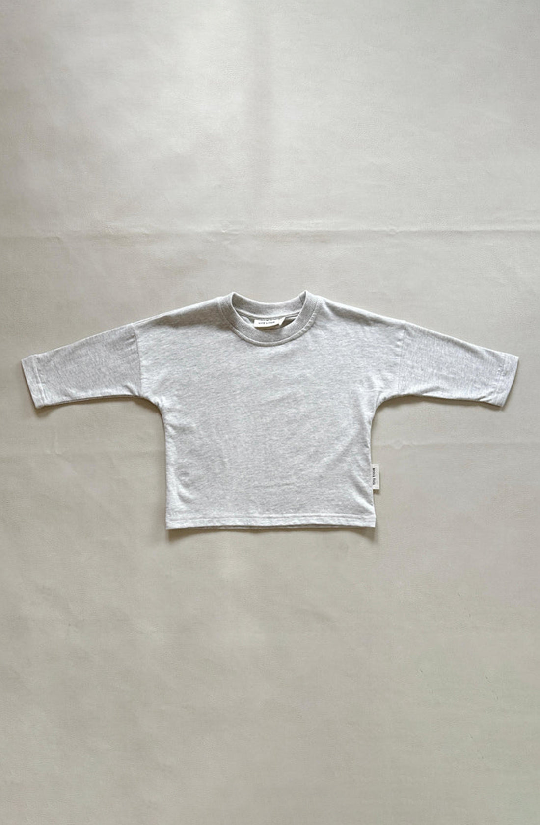 The Romy Long Sleeve Tee Light Grey Marle from TINY TROVE is laid flat on a smooth white surface. With drop shoulders and a relaxed fit, this 100% cotton shirt features a crew neckline and appears to be made from soft fabric. No visible designs or logos are present.