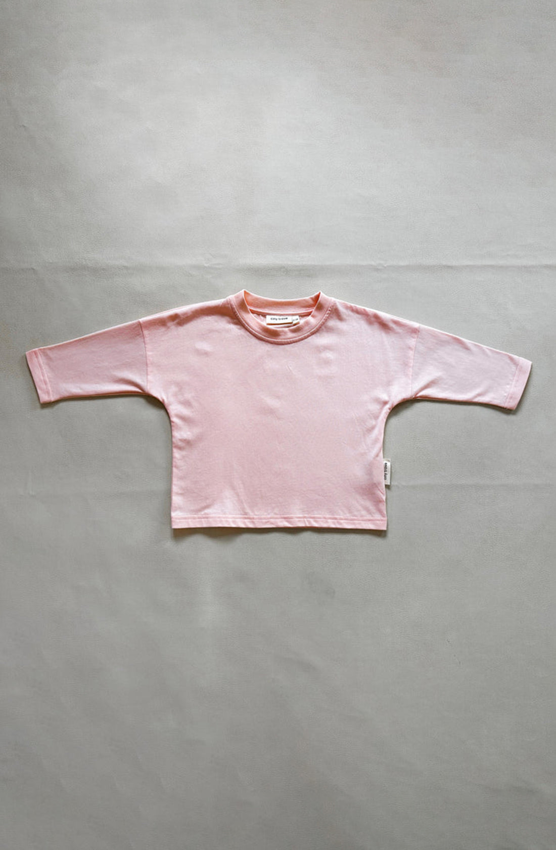 The Romy Long Sleeve Tee Pink by TINY TROVE is laid flat on a neutral-colored surface. Made of 100% cotton, the shirt features a soft, comfortable fabric with a round neckline. It also has drop shoulders for a relaxed fit and includes a small tag at the side seam.