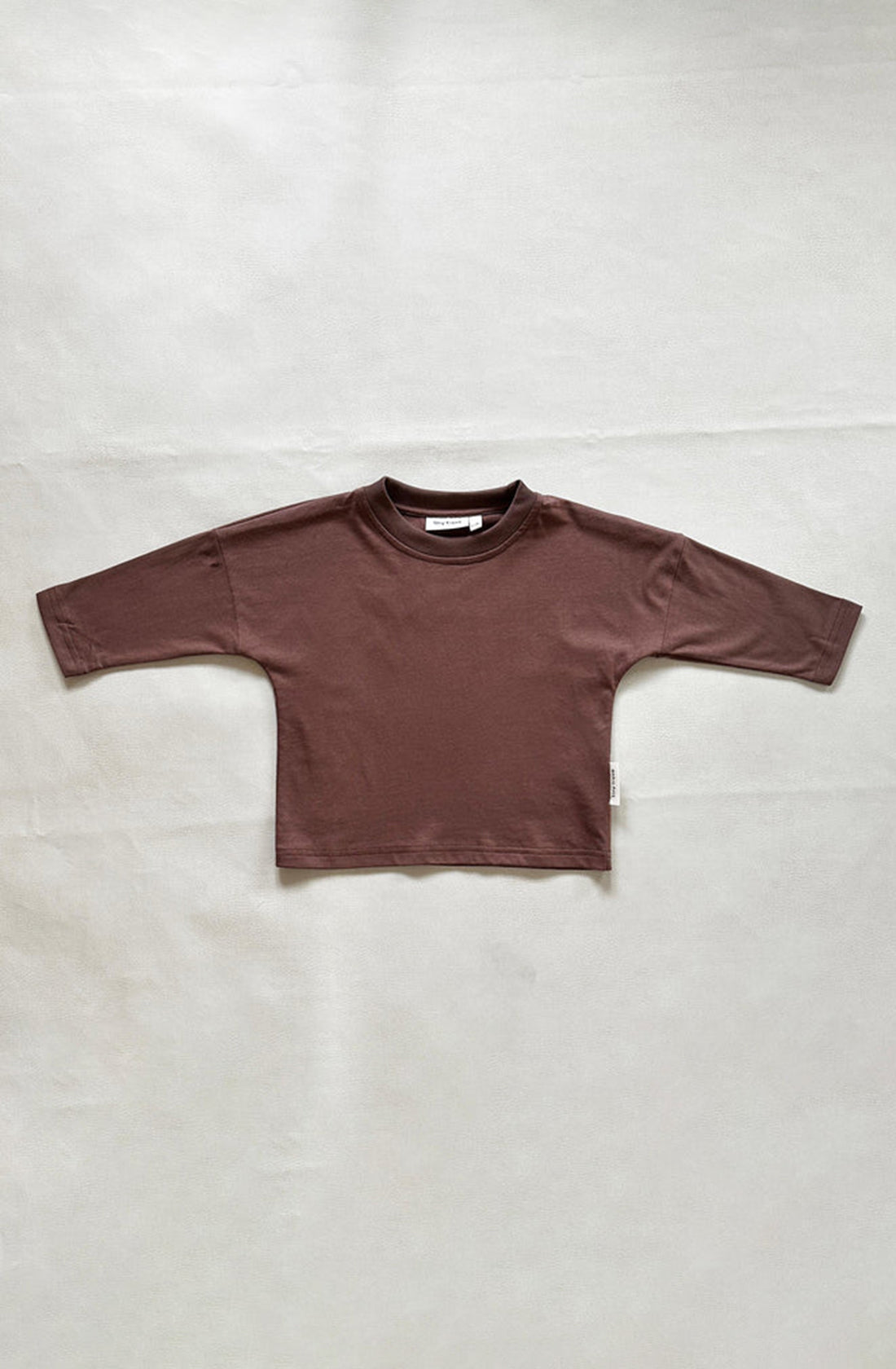 The Romy Long Sleeve Tee Mocha from TINY TROVE lies flat on a light gray surface. Made from 100% cotton, this brown baby shirt features a round neckline and visible stitching along the hem and sleeves. Designed for comfort, it boasts drop shoulders and a relaxed fit. A small white tag is visible on the side seam.