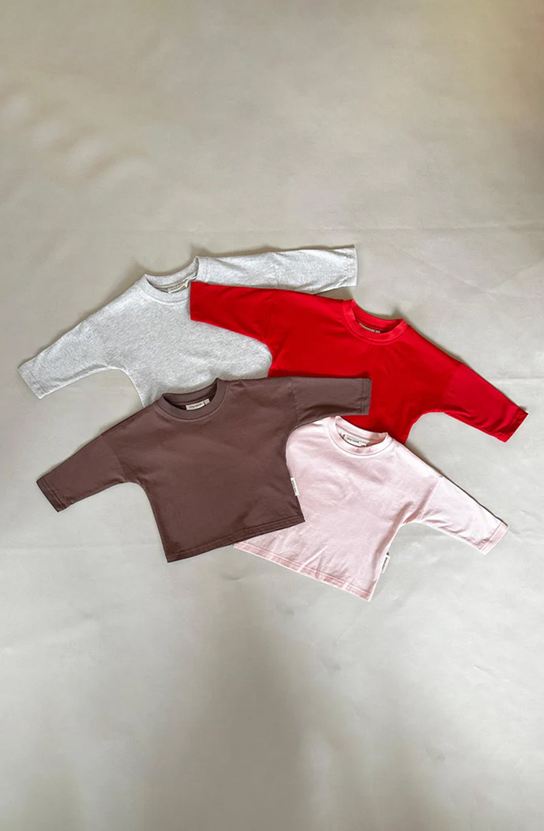 Four TINY TROVE Romy Long Sleeve Tees are laid out against a neutral background. From top to bottom, the relaxed fit shirts are light grey marle, red, brown, and light pink in color. Each shirt is plain with no visible designs or logos, perfect for a simple yet stylish look.