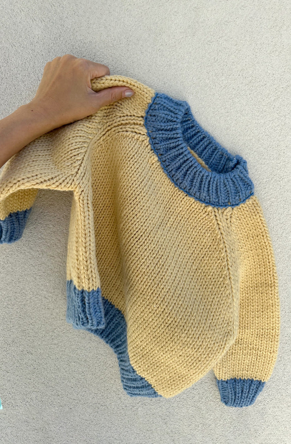 A hand holding the TINY TROVE Willa Contrast Knit Cream/Blue sweater, featuring a chunky knit design with sky blue accents on the collar, cuffs, and bottom hem. The texture and stitches of the wool blend sweater are clearly visible against a light background.