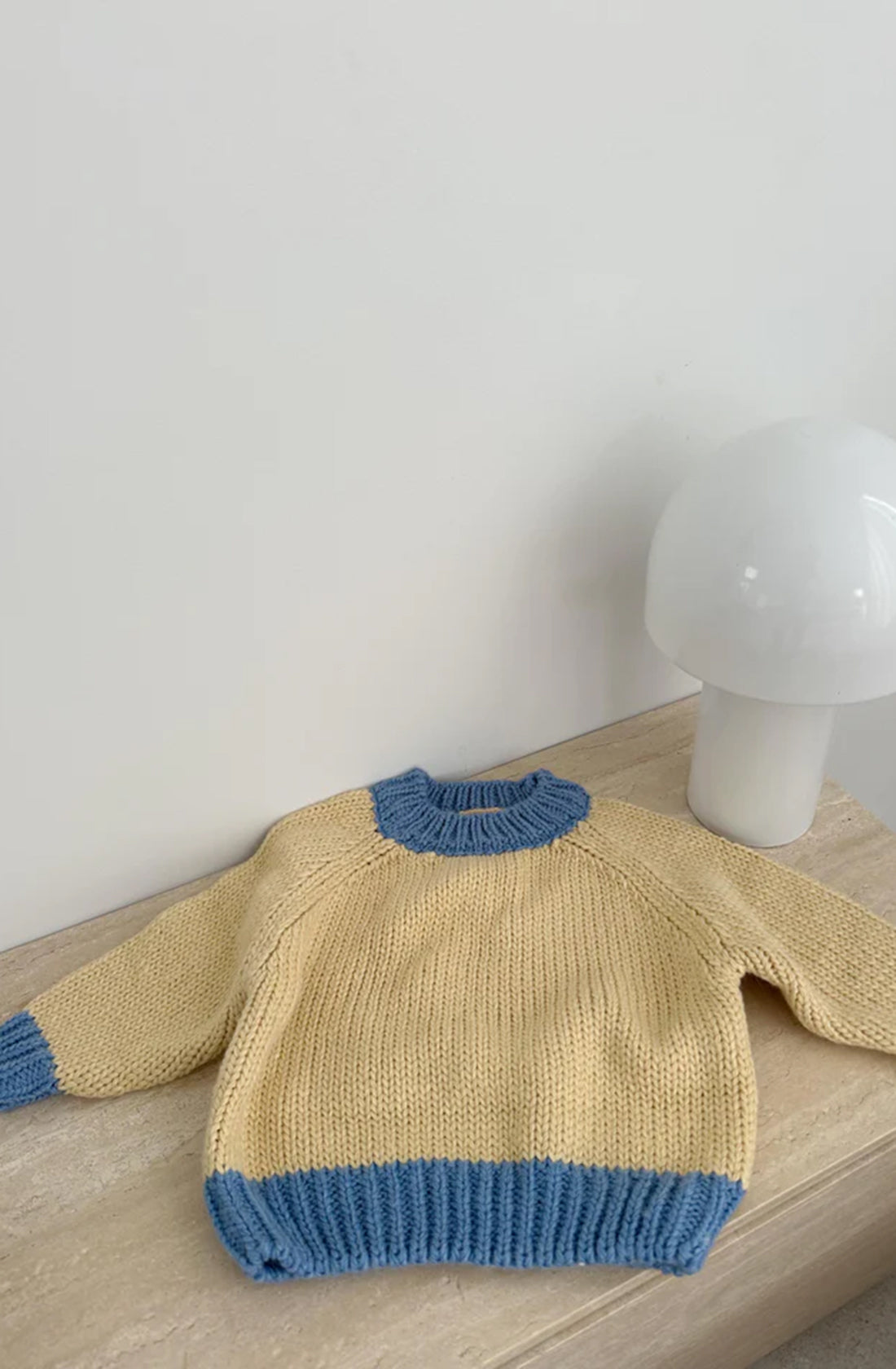 The Willa Contrast Knit Cream/Blue sweater by TINY TROVE, featuring an oversized fit with blue cuffs, collar, and hem, is laid out neatly on a wooden surface. To the right of the sweater, there is a white modern lamp with a rounded shape resembling a mushroom. The background features a plain white wall.