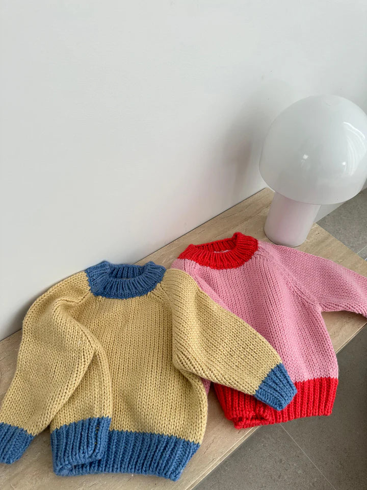 Two chunky knit children's sweaters from TINY TROVE are displayed on a light wooden surface. One sweater, made from recycled acrylic, is yellow with blue cuffs, collar, and hem; the other is the Willa Contrast Knit in pink with red trims. Both have an oversized fit. A white, dome-shaped lamp is placed behind them against a plain wall.