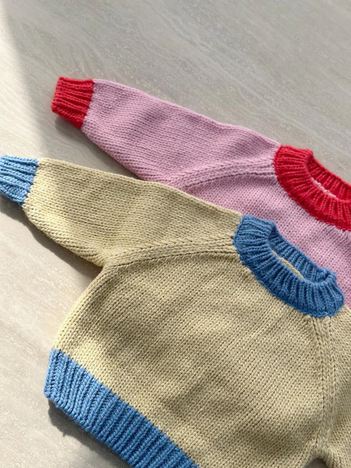 Two hand-knitted baby sweaters are laid flat on a light surface. The top sweater, featuring an oversized fit, is pink with red cuffs and neckline, while the bottom sweater is the TINY TROVE Willa Contrast Knit Cream/Blue. Sunlight casts soft shadows across the cozy wool blend sweaters.