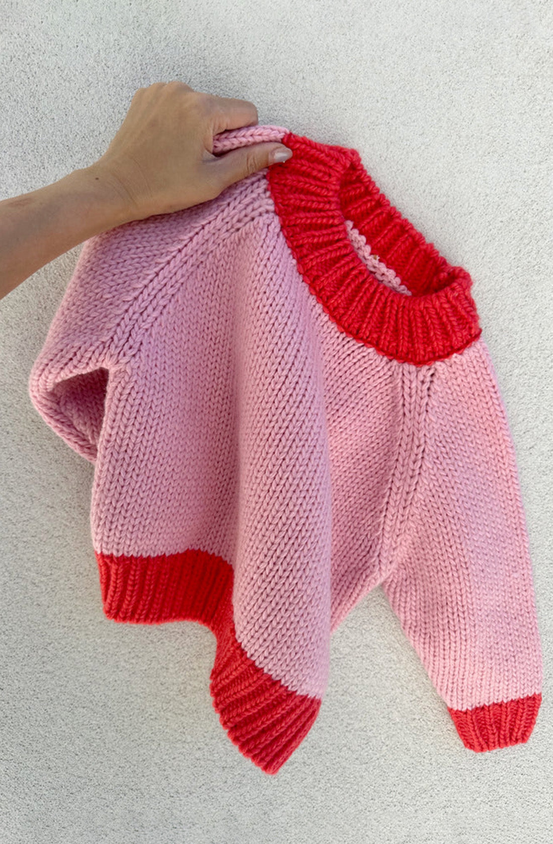 A hand is holding up the TINY TROVE "Willa Contrast Knit Pink/Red" sweater against a light background. This pink knitted sweater features red ribbed cuffs, hem, and collar. It boasts an oversized fit and is made from recycled acrylic for an eco-friendly choice.