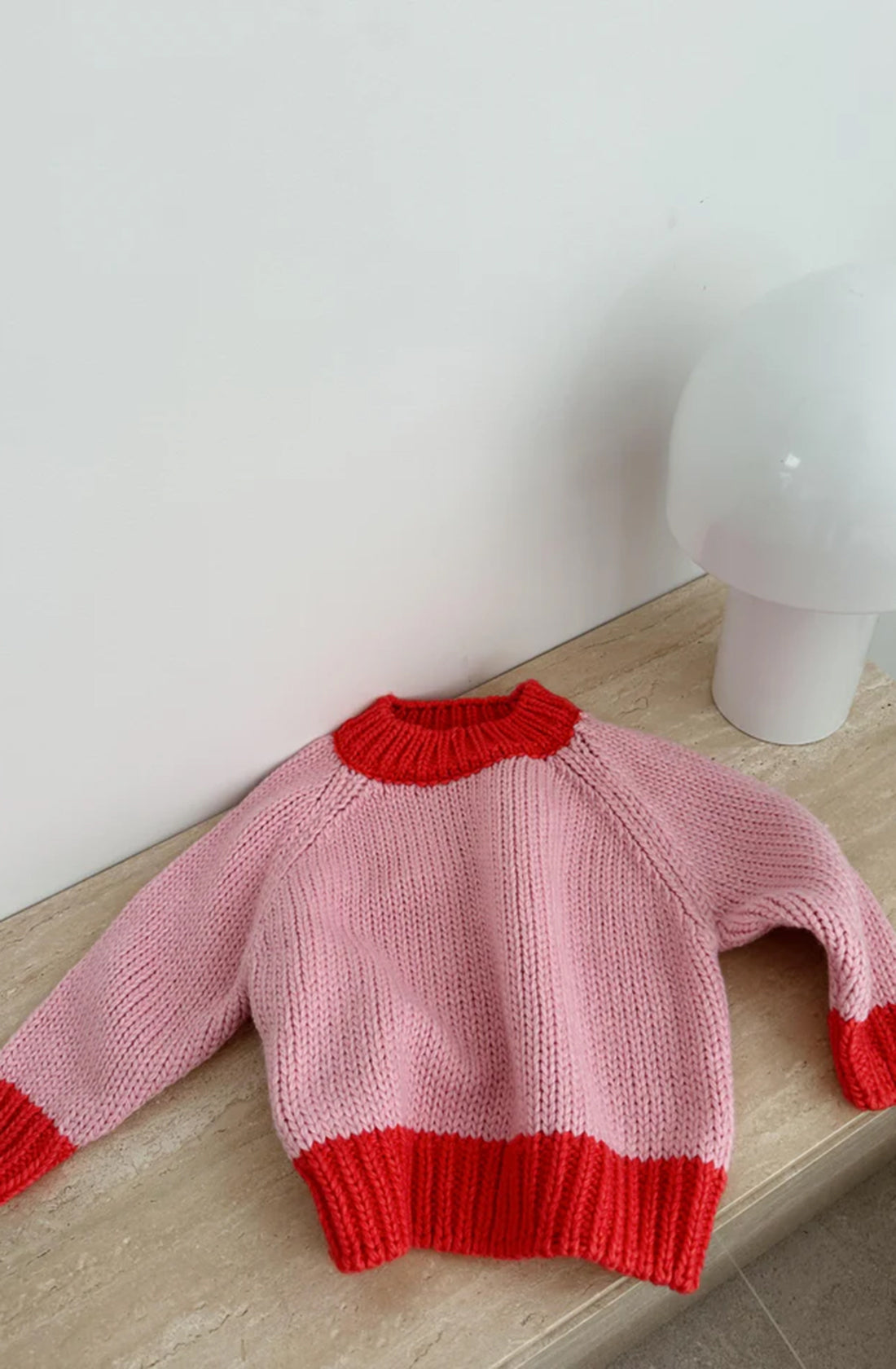 The Willa Contrast Knit Pink/Red sweater by TINY TROVE, featuring chunky knit in pink with red accents on the collar, cuffs, and hem, is laid flat on a wooden surface. Positioned to its right against a plain white wall is a white lamp with a round, smooth base.