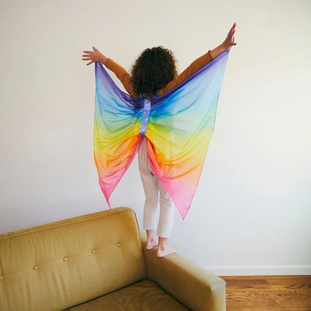 A child with curly hair stands on a couch, arms outstretched, wearing SARAH'S SILKS' vibrant Rainbow Wings for dress-up fun. The background showcases a plain white wall and a wooden floor.