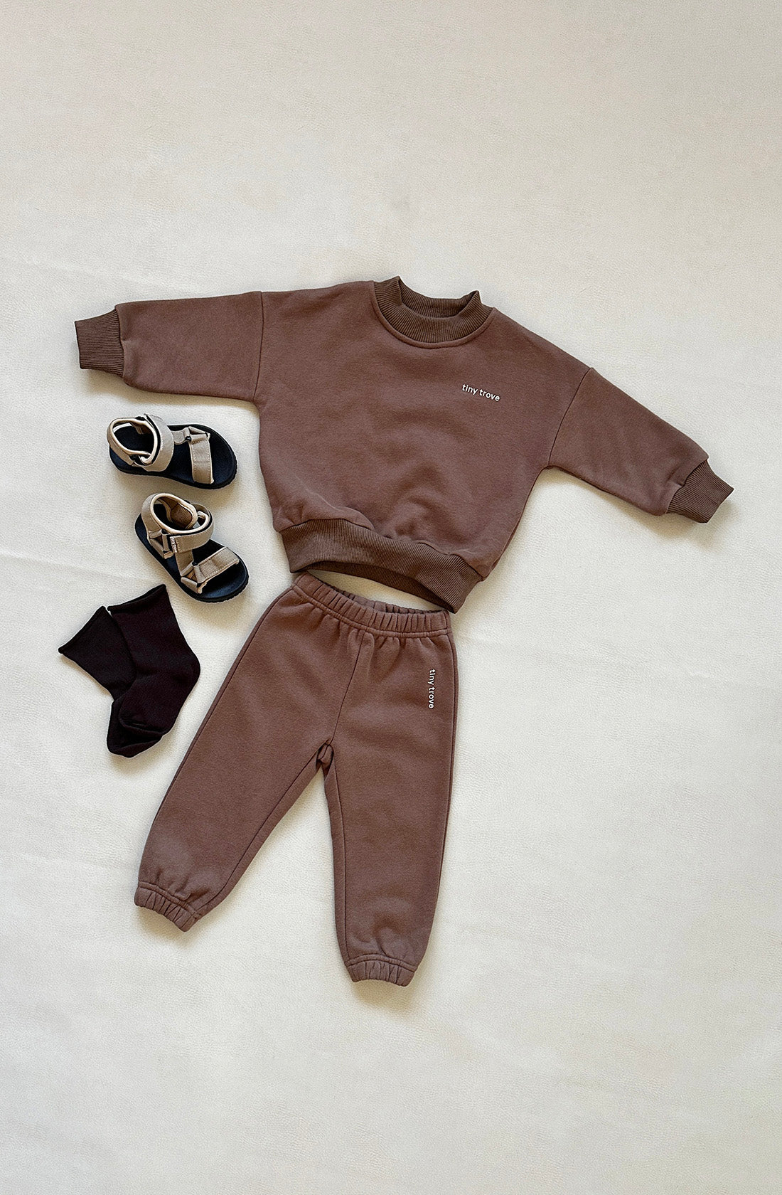 Showcased is the Woodie 3D Logo Tracksuit Mocha by TINY TROVE, which includes a mocha sweatshirt and matching fleece cotton sweatpants. Adjacent to it are a pair of black socks and sandals featuring black and beige straps, all elegantly arranged against a soft beige backdrop.