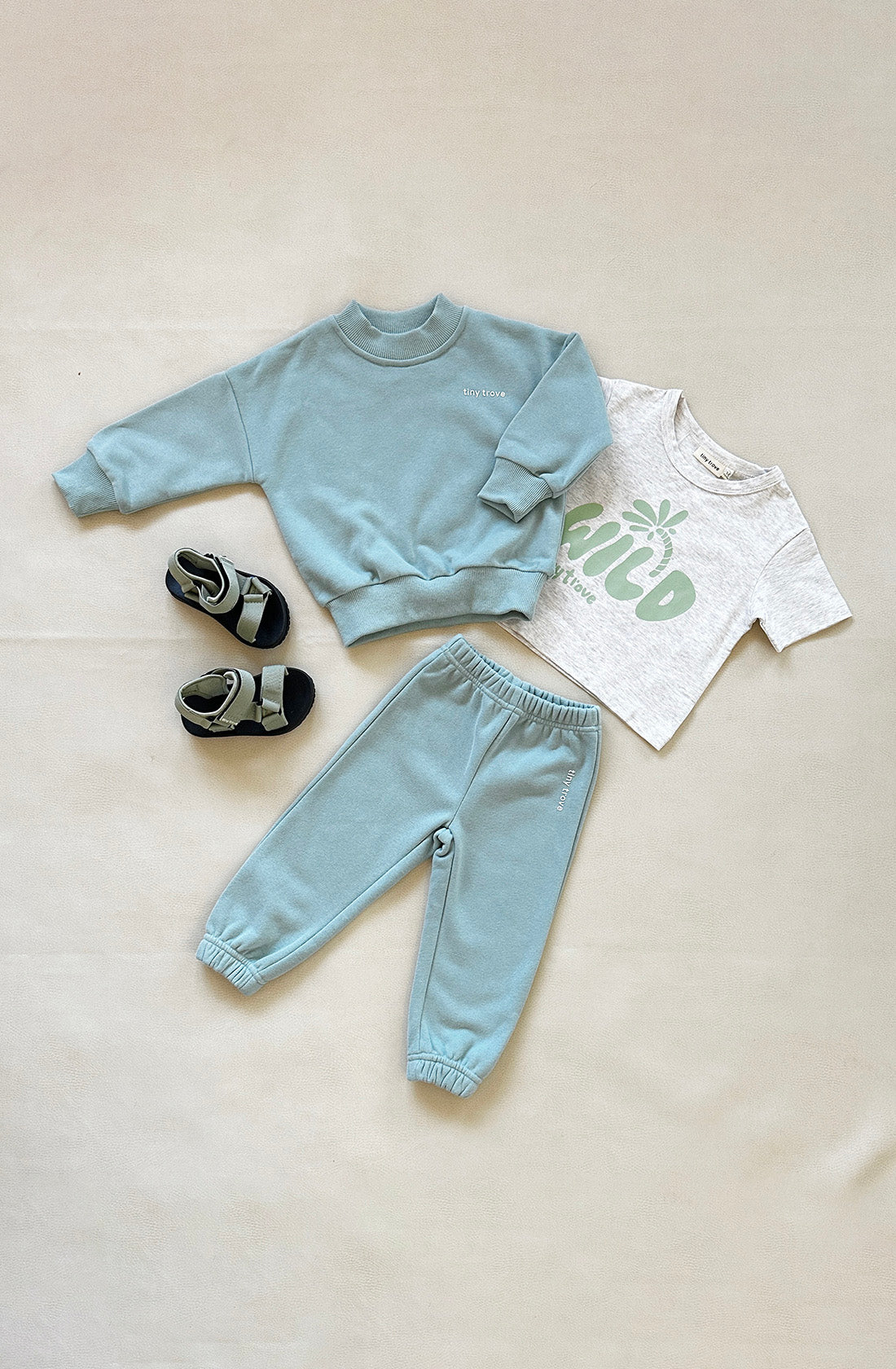 A neatly arranged toddler outfit set from TINY TROVE, named the Woodie 3D Logo Tracksuit in Seafoam, lies on a light beige background. The soft cotton jersey set includes a light blue sweatshirt, matching jogger pants, a white t-shirt featuring a green "kidd" 3D logo and palm tree graphic, and a pair of gray sandals with black soles.