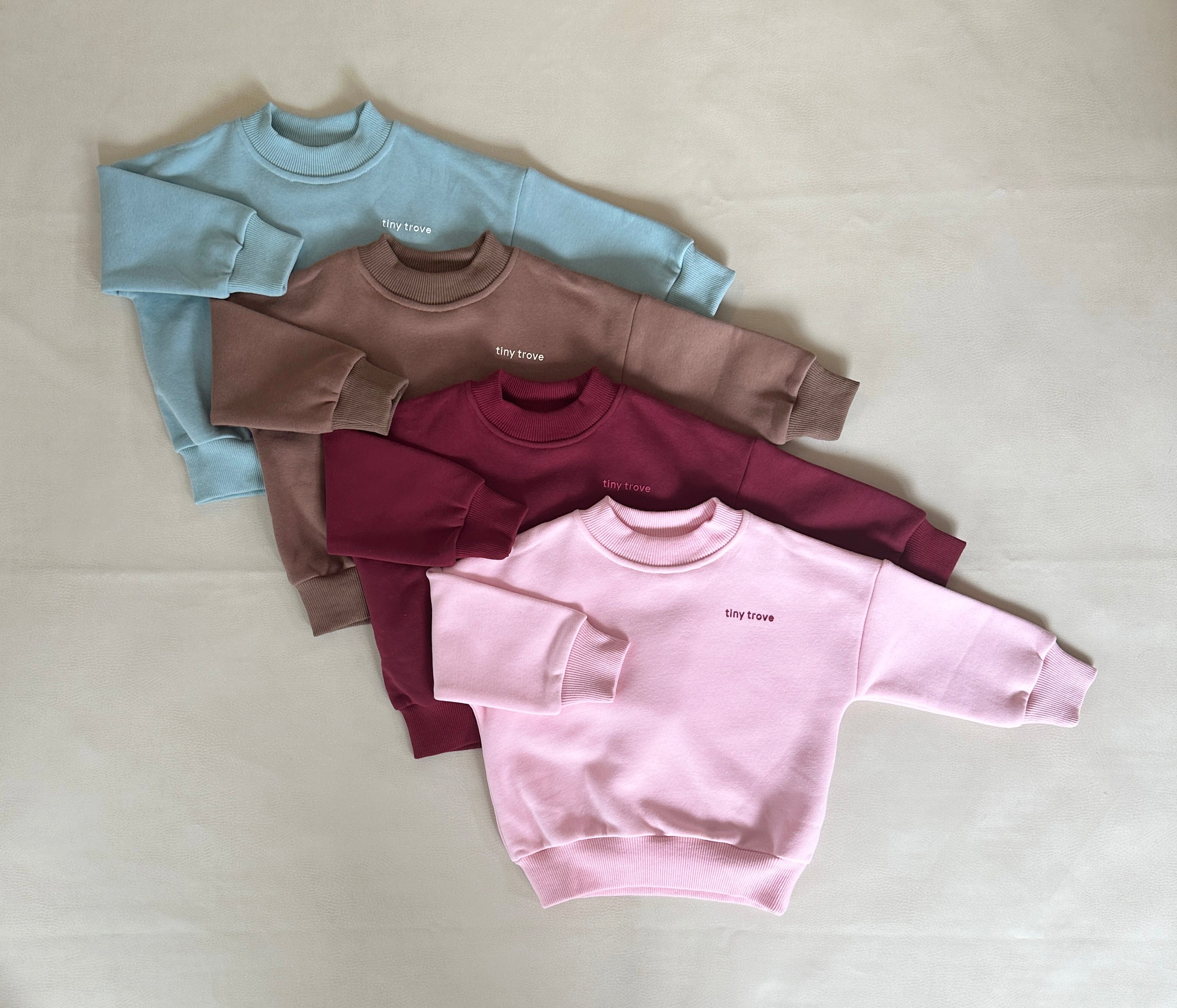 Four children's crewneck sweaters, crafted from soft cotton jersey, are arranged in a fanned display. From top to bottom, the colors are light blue, light brown, maroon, and light pink. Each sweater features the words "Stay brave" embroidered on the chest area. The product is part of the Woodie 3D Logo Tracksuit Mocha collection by TINY TROVE.
