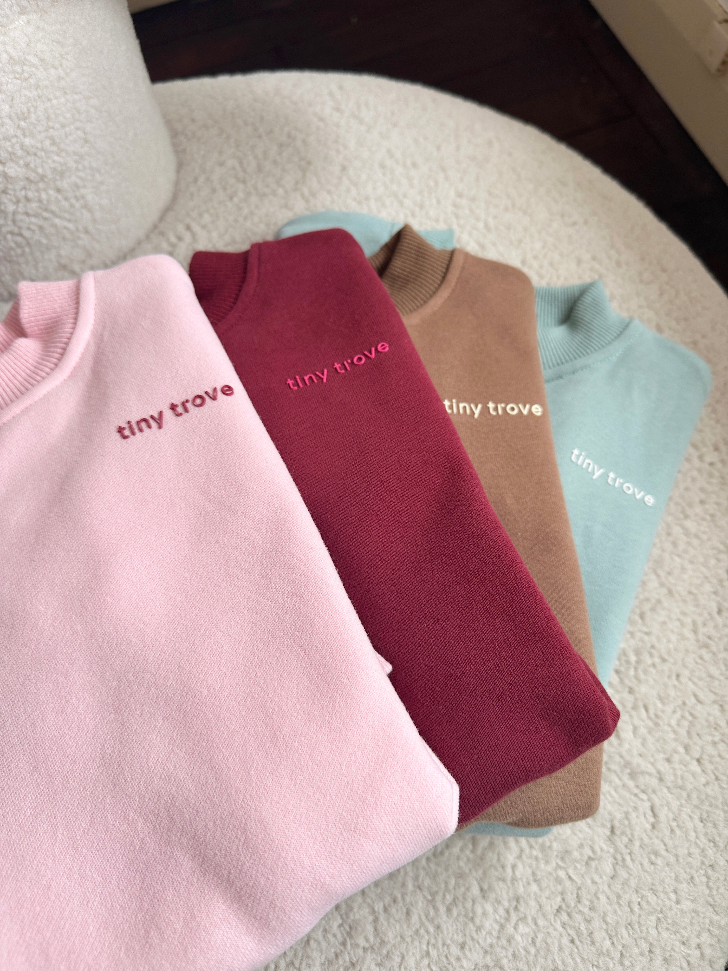 Four folded Woodie 3D Logo Tracksuits in mocha from TINY TROVE are displayed on a light, textured surface. The tracksuits, crafted from soft cotton jersey, come in pastel pink, deep red, camel brown, and pastel blue. Each one features "tiny trove" embroidered on the chest in matching or contrasting colors.