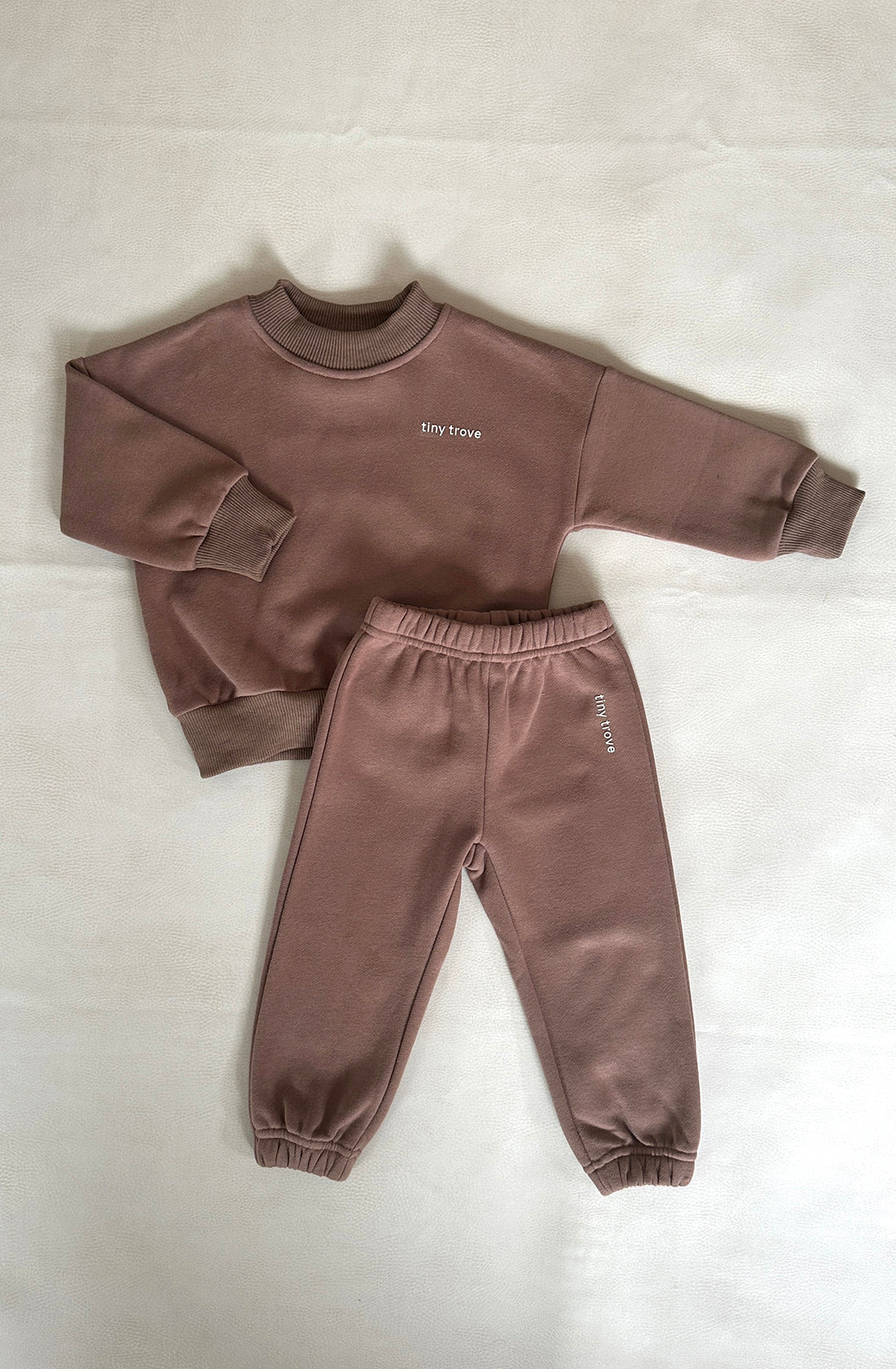 A children's outfit, known as the Woodie 3D Logo Tracksuit Mocha from TINY TROVE, is displayed on a light-colored surface. This ensemble includes a matching brown sweatshirt and jogger pants, both crafted from soft cotton jersey. The sweatshirt showcases a stylish "tiny trove" 3D logo on the chest, while the jogger pants feature the same text on the upper left leg. Both pieces are designed with ribbed cuffs and waistbands for added comfort.