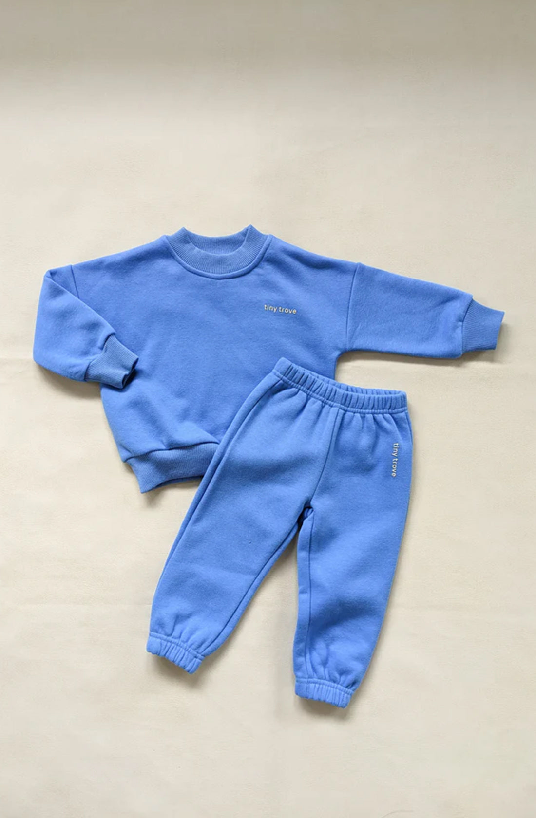 A matching set of children's clothing laid flat on a beige background. The comfortable wear includes the Woodie 3D Logo Tracksuit Ocean by TINY TROVE, featuring a blue long-sleeve sweatshirt with "tiny love" written in 3D on the chest and a pair of fleece cotton blue sweatpants with "tiny love" on the left thigh.