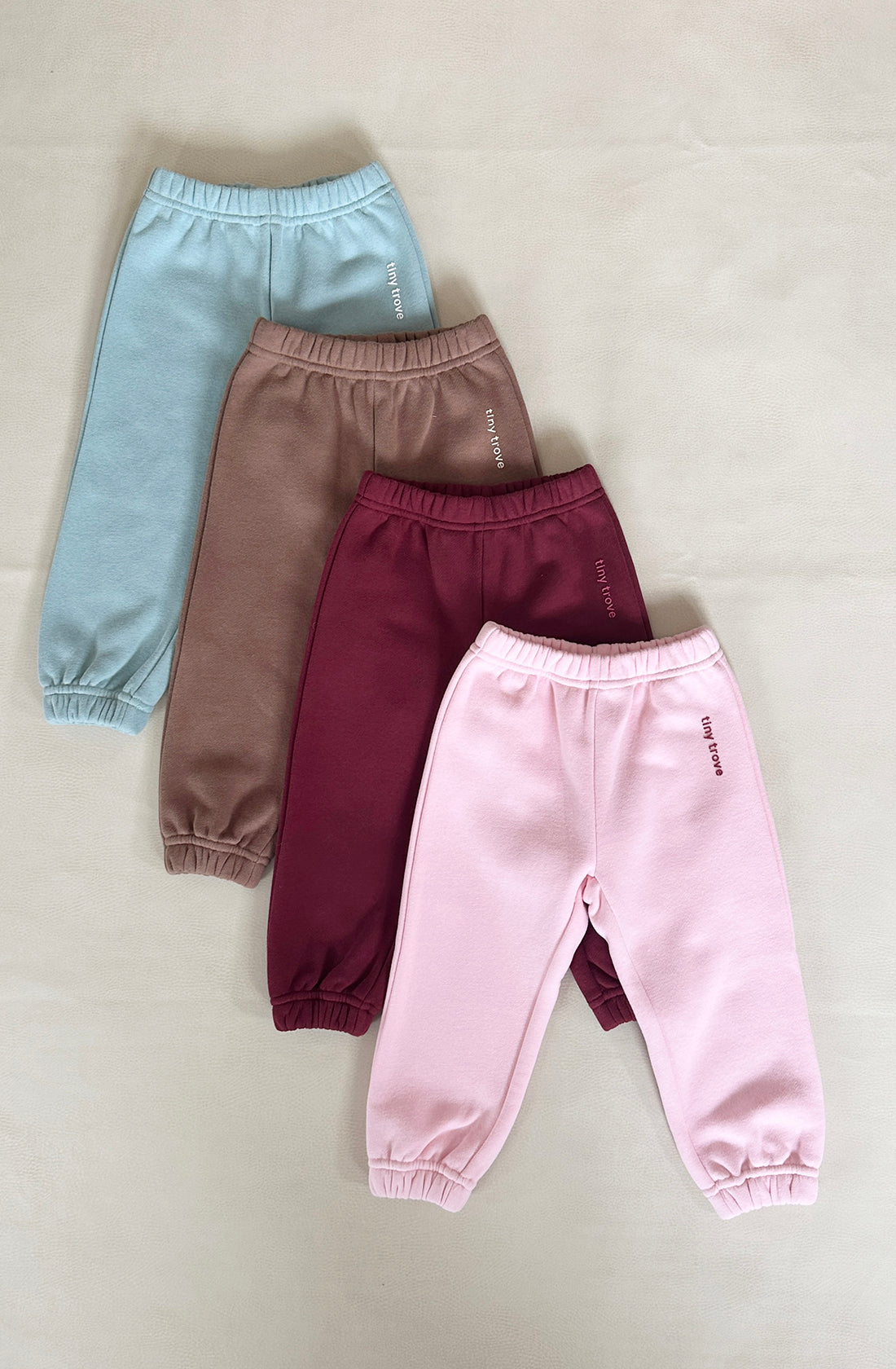 Four pairs of the TINY TROVE Woodie 3D Logo Tracksuit in Mocha — light blue, brown, maroon, and pink — are arranged in a line on a flat surface. Each pair is made of soft cotton jersey with elastic cuffs at the ankles and features a small printed text on the upper left leg.