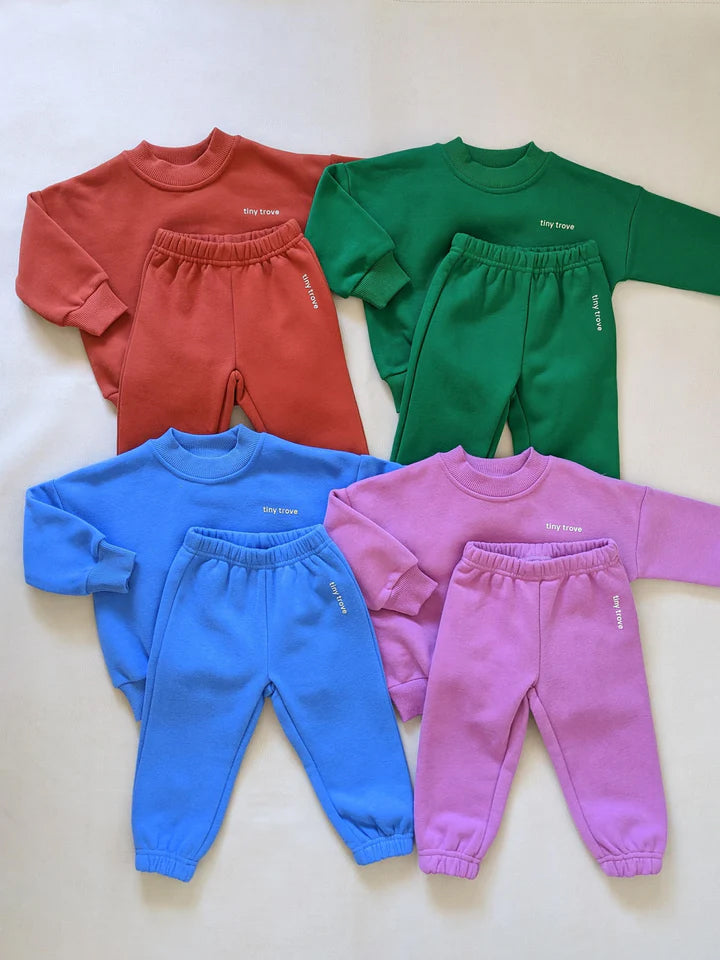 Five sets of children's tracksuits in various colors are displayed on a flat surface. The Woodie 3D Logo Tracksuit Ocean by TINY TROVE includes a long-sleeved top and matching pants available in red, green, light blue, dark blue, and purple. Each set features the "tiny crew" branding with a 3D logo on both the top and pants.