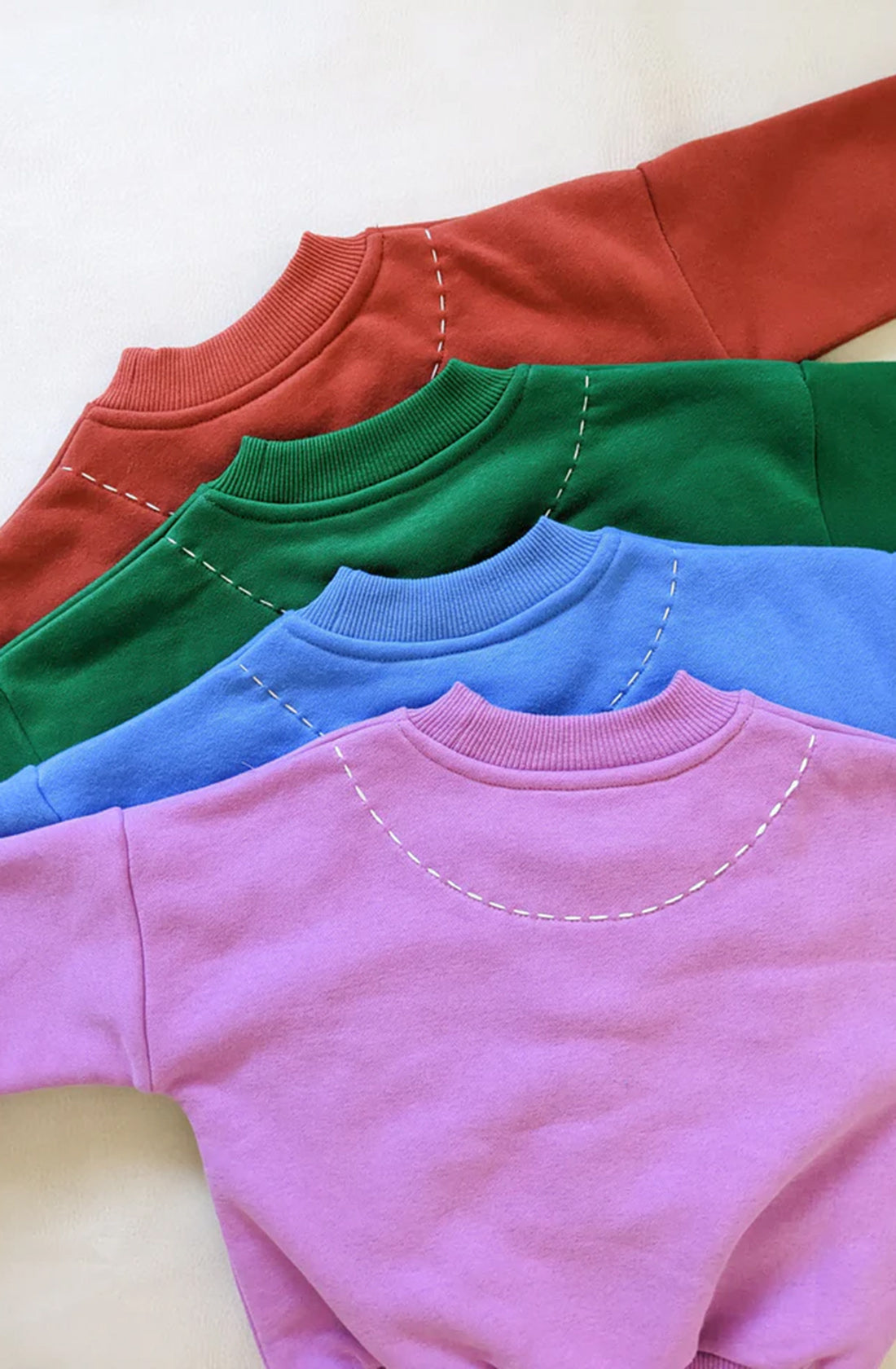 Four colorful sweaters with contrast stitch accents, made of comfortable fleece cotton, are neatly stacked. From top to bottom, the sweaters are red, green, blue, and pink. Each sweater features a visible stitch pattern around the neckline. The background is off-white.

Product:
  Product Name: Woodie 3D Logo Tracksuit Ocean
  Brand Name: TINY TROVE