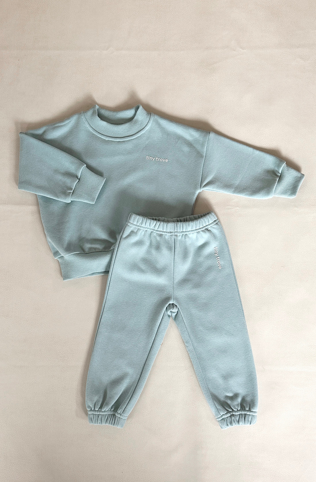 A seafoam-colored Woodie 3D Logo Tracksuit in toddler size, made from soft cotton jersey, is neatly laid out on a cream-colored surface. The set includes a long-sleeved sweatshirt and matching sweatpants, both featuring ribbed cuffs and hems. The words "TINY TROVE" are embroidered in white on the chest of the sweatshirt and the leg of the pants.