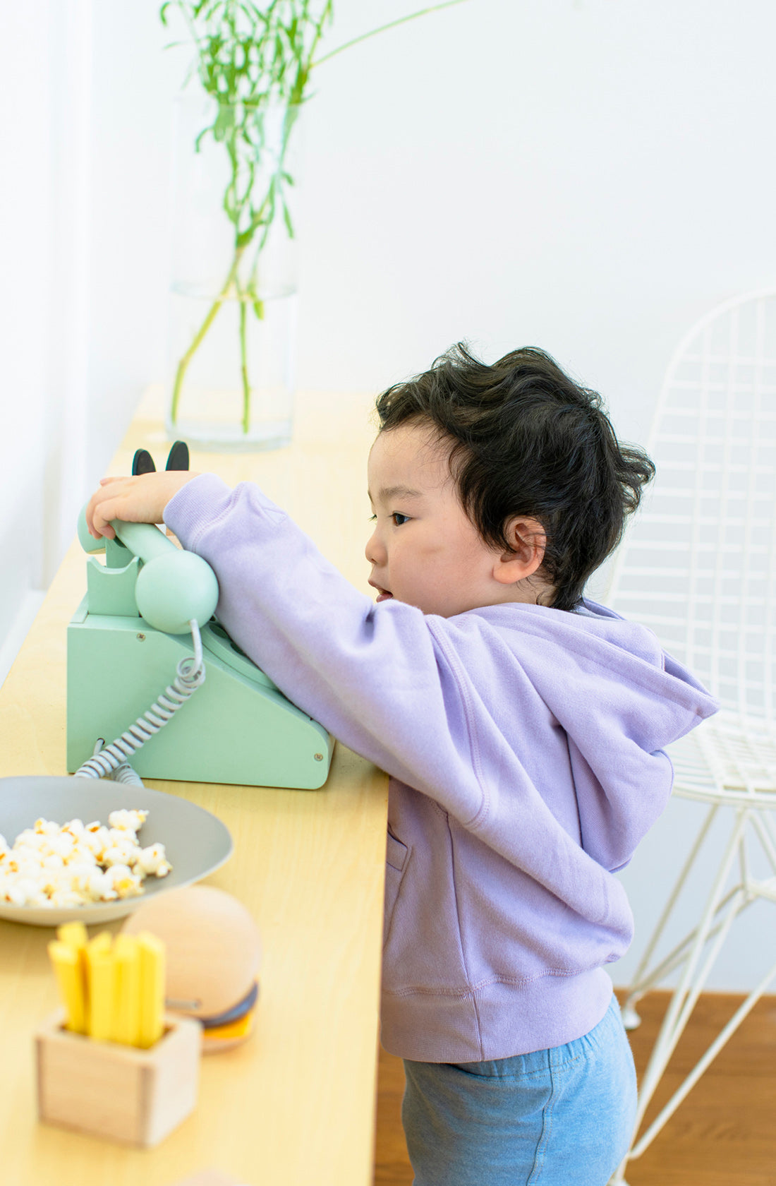 A young child wearing a lavender hoodie stands on tiptoes to reach the KIKO & GG Retro Telephone Green, their imagination turning it into a playful toy. They hold the receiver while standing by a wooden table with snacks, including popcorn and cheese sticks. A white wire chair and a plant are in the background.