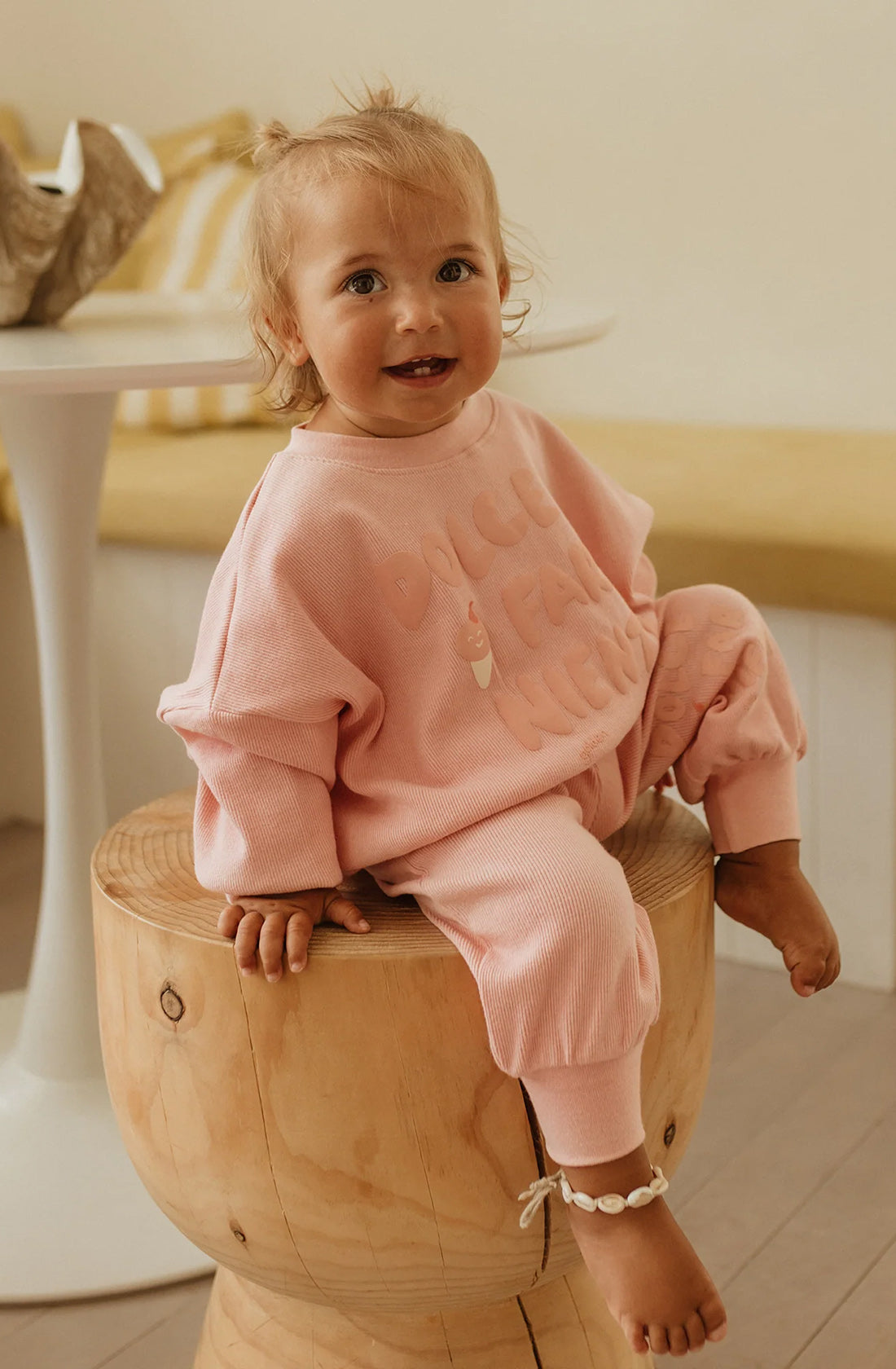 A toddler sits on a round, wooden stool. The child is wearing light pink, cozy Dolce Lounge Pant Pink Lemonade by GOLDEN CHILDREN with a relaxed fit and is smiling. There's a white table and a yellow cushion in the background. The toddler has a light bracelet on one ankle.