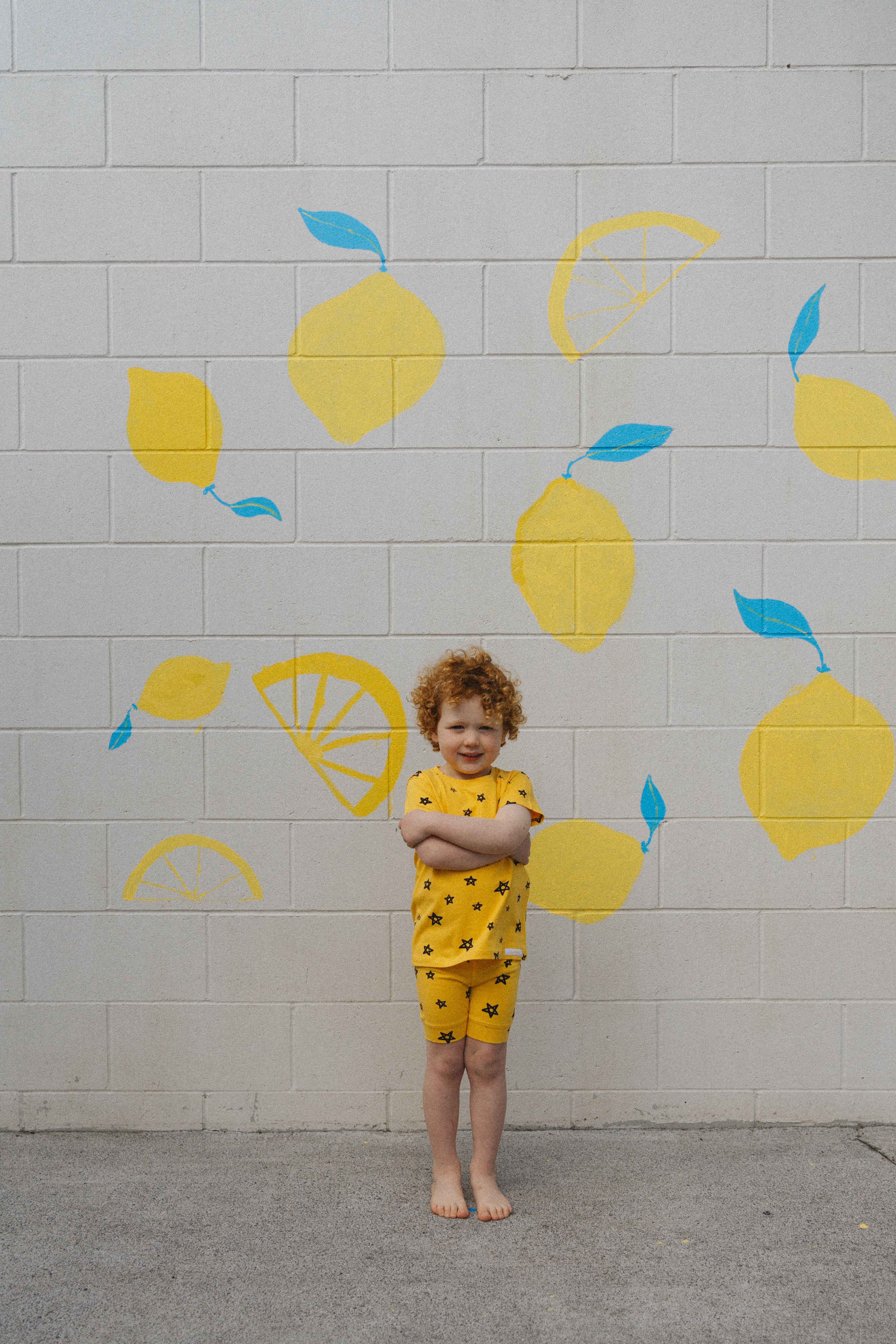 A child with curly hair stands barefoot against a white brick wall decorated with yellow lemons and blue leaves, wearing the Star Shortie PJ Set Lemon from G.NANCY. Their charming matching yellow outfit, featuring a star pattern, showcases the ultimate comfort fit as they cross their arms.