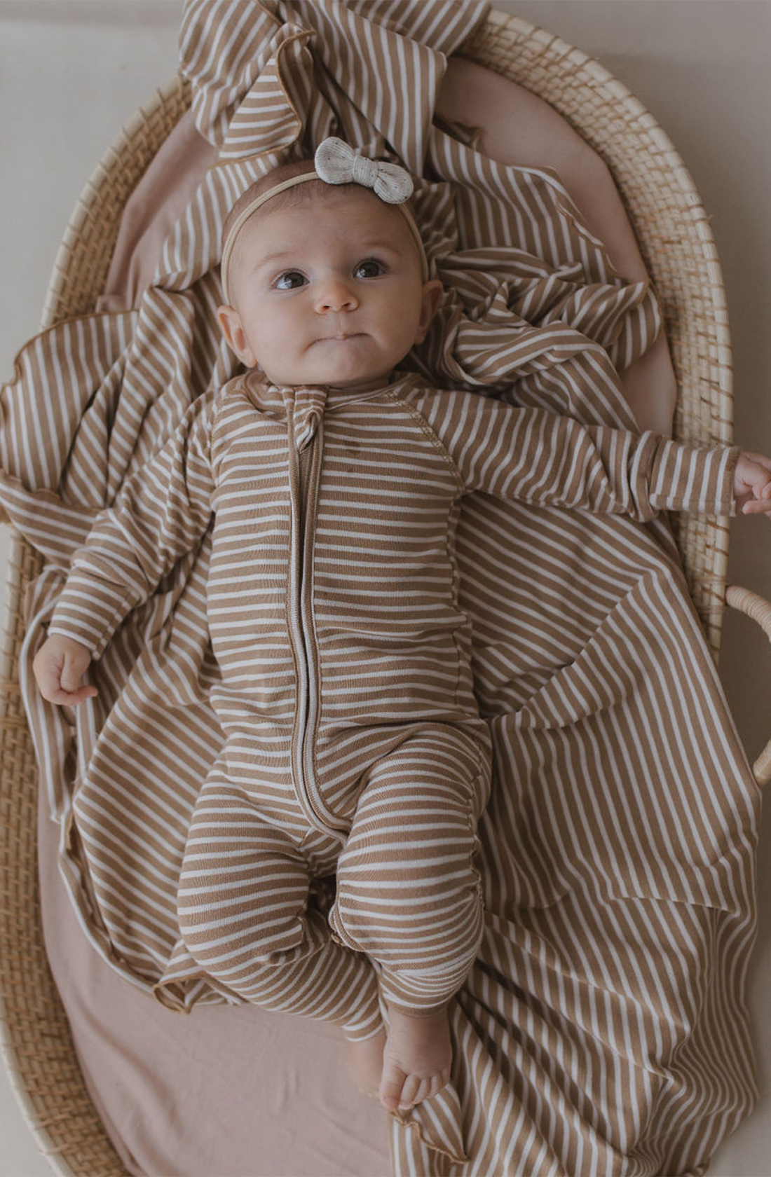 A baby wearing SUSUKOSHI's brown and white striped Zip LS Pants Autumn Stripe made from GOTS Organic Cotton lies in a basket lined with matching fabric. The baby has a headband with a bow and is looking upwards with a curious expression.