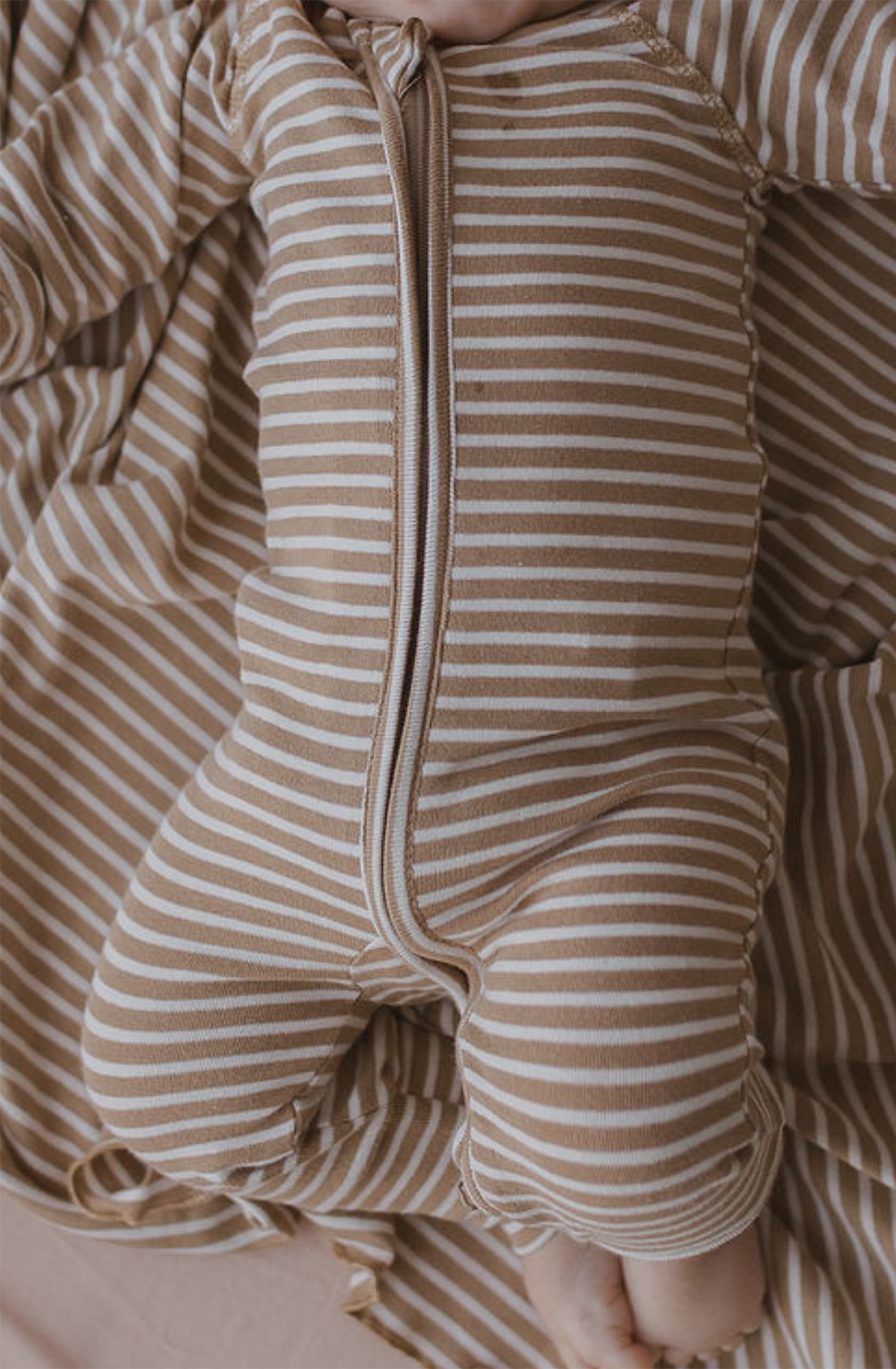 A baby dressed in a brown and white striped SUSUKOSHI Zip LS Pants Autumn Stripe made from GOTS Organic Cotton is lying on a matching striped blanket. The baby's legs are slightly bent, and the outfit features a two-way zip running down the front. Only part of the baby’s face is visible in the upper right corner.