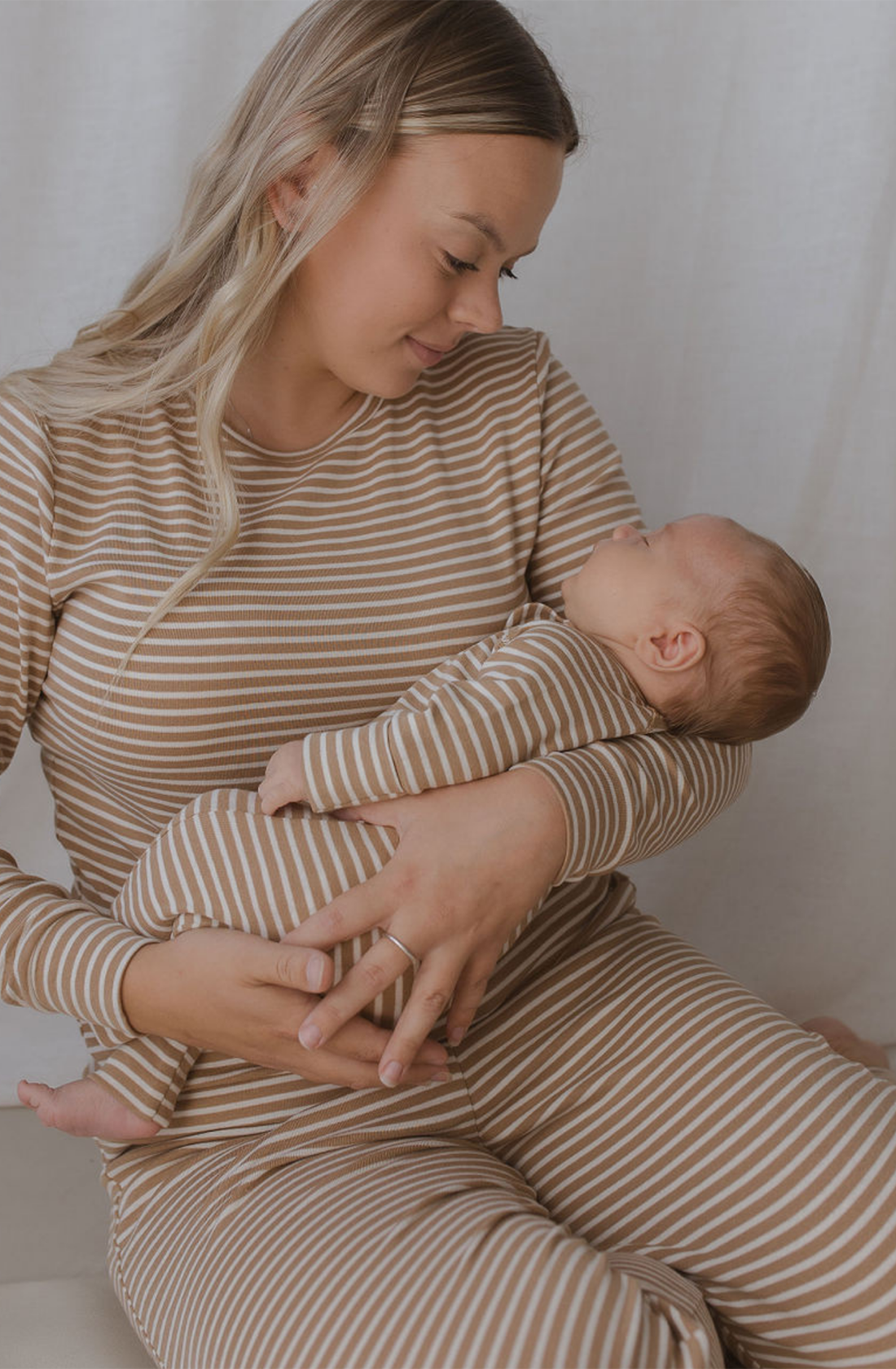 A woman cradles a baby in her arms, both wearing matching SUSUKOSHI Zip LS Pants Autumn Stripe made from GOTS Organic Cotton. The woman has long blonde hair and looks lovingly down at the baby, who is resting comfortably against her chest. The background is a plain light color.