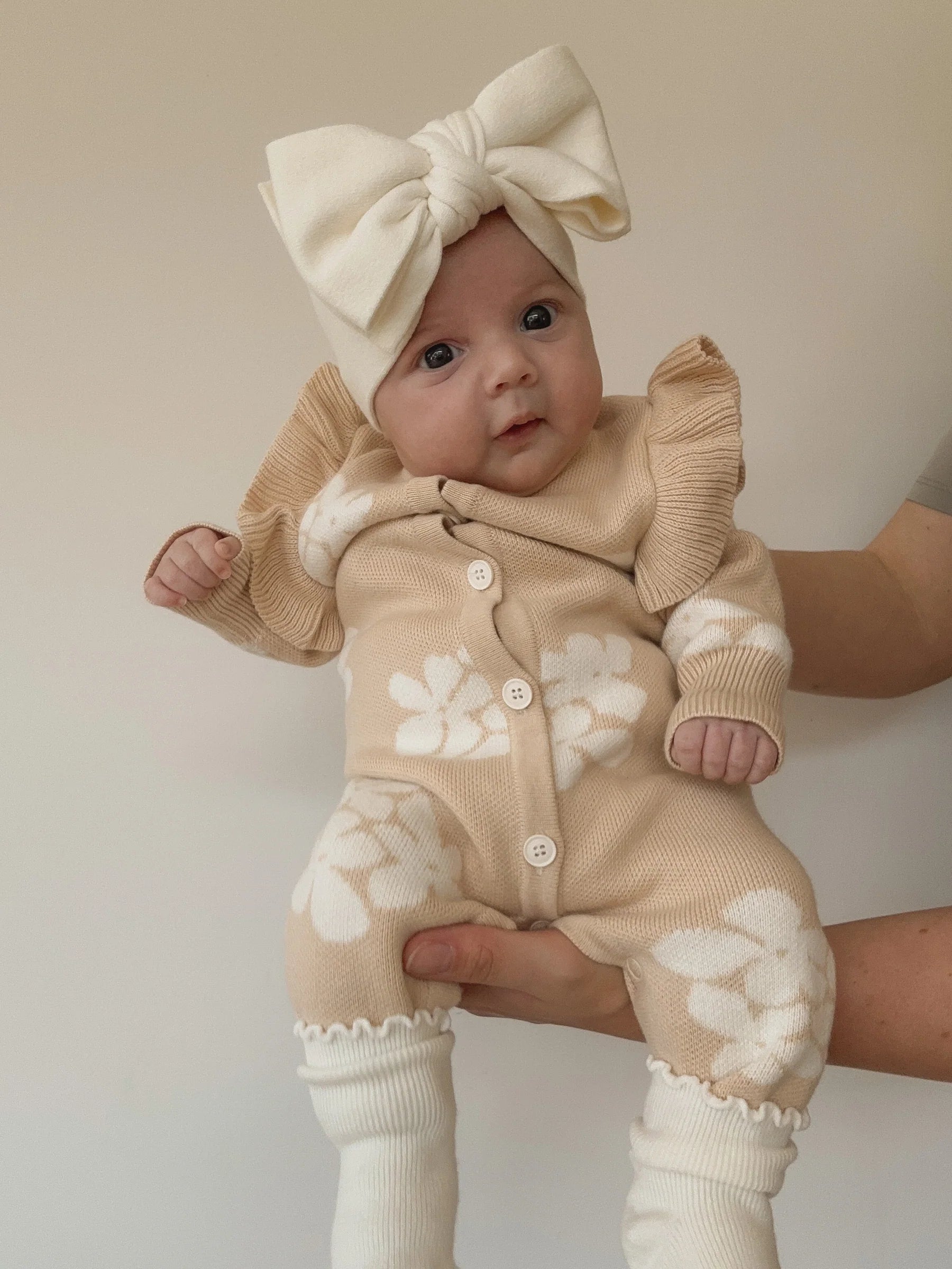 A baby wears a beige outfit with white floral patterns and the Ziggy Lou - Headband Milk by ZIGGY LOU, held safely by an adult’s hand against a plain light background.