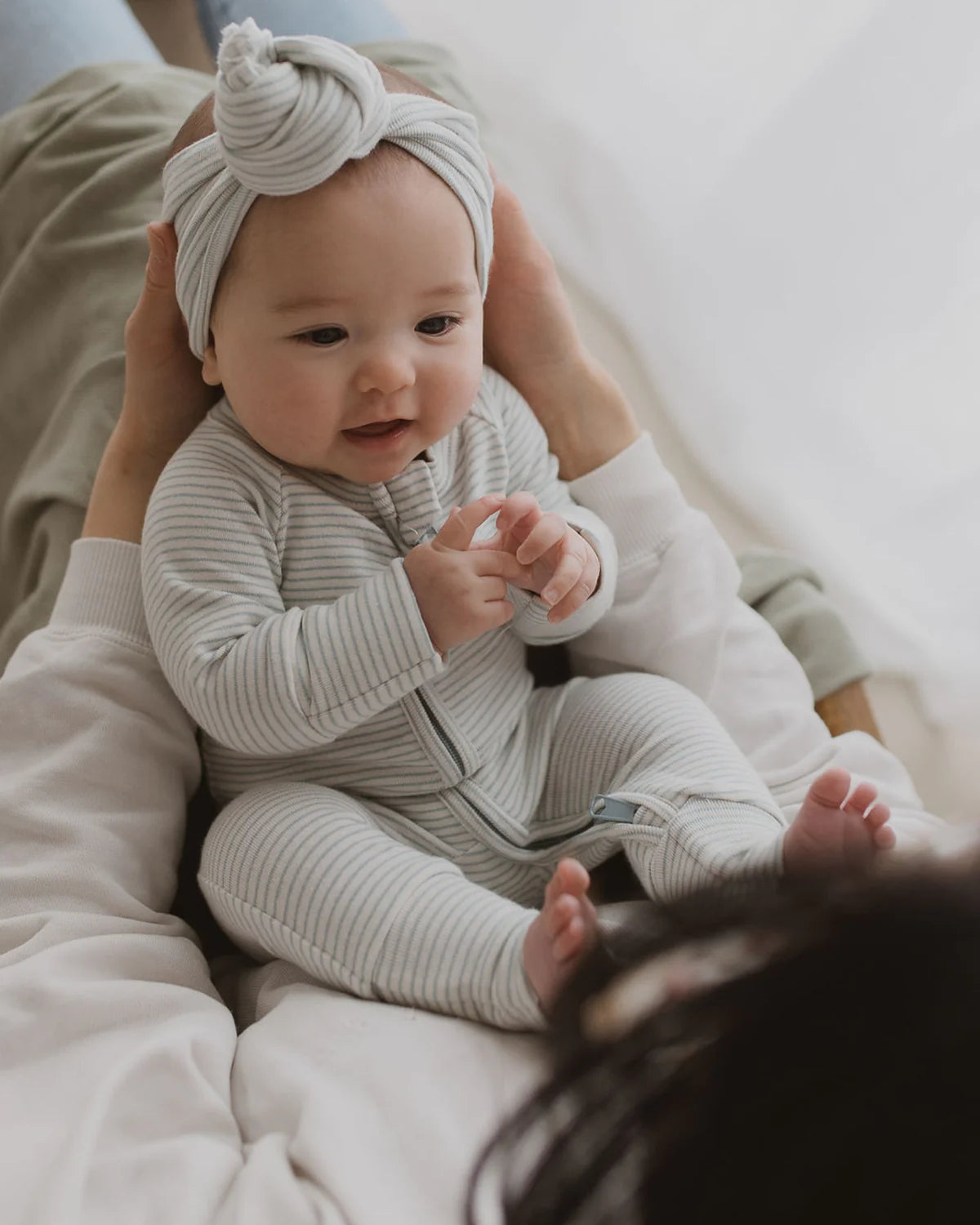 A baby wearing the SUSUKOSHI Zip Suit Seashore and a turban-style headband sits on an adult's lap. The baby is smiling and looking forward, with the adult's hands gently supporting them from behind. The background is softly lit, creating a warm and cozy atmosphere.