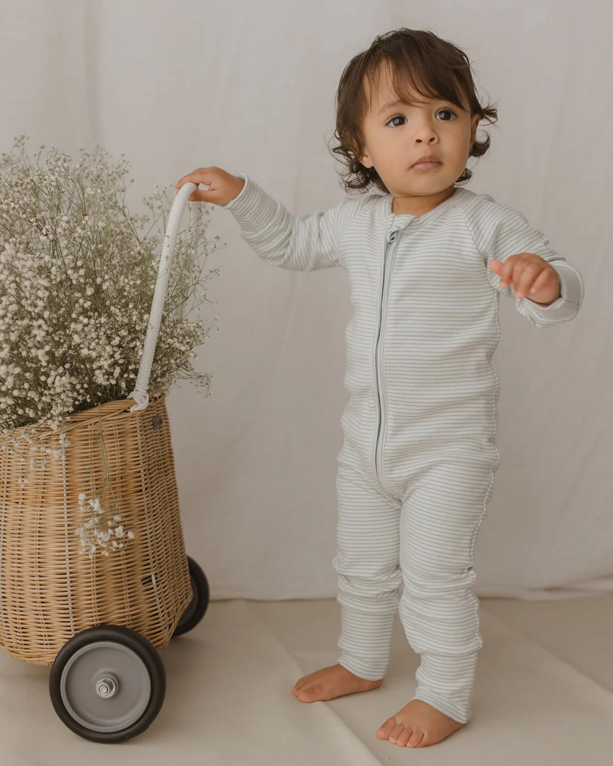 A toddler wearing the SUSUKOSHI Zip Suit Seashore stands next to a wicker basket on wheels filled with white flowers. The child, dressed in this organic cotton baby zip suit, is holding the basket's handle and looking into the distance with a calm expression against a background of neutral, soft fabric.