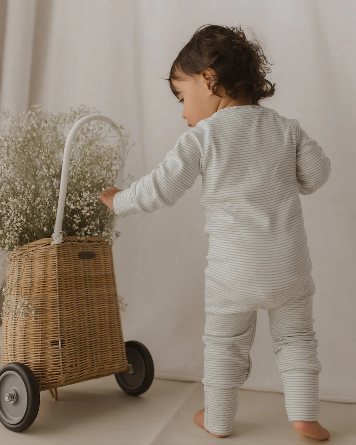 A toddler wearing a SUSUKOSHI Zip Suit Seashore, made of light grey, striped organic cotton, is standing and facing away from the camera. The child is interacting with a wicker basket on wheels that is filled with delicate white flowers. The background is softly lit and neutral-colored.