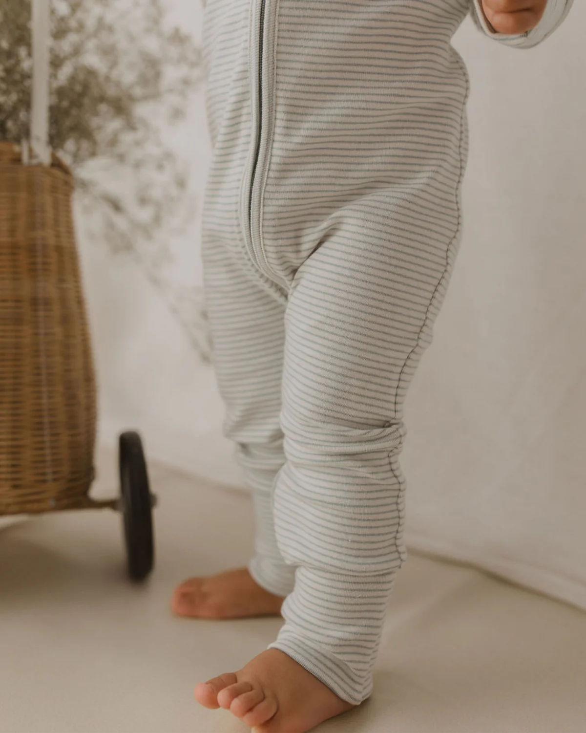 A baby, dressed in a gray and white striped SUSUKOSHI Zip Suit Seashore made of organic cotton, stands barefoot on a beige surface. The image focuses on the lower body, capturing the legs and feet. A wicker basket is partly visible in the background on the left.