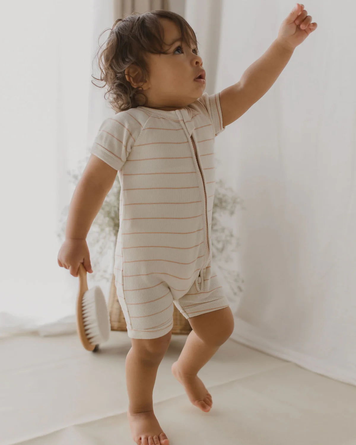 A toddler stands barefoot on a light-colored floor, holding a brush in one hand and reaching upward with the other. The child wears a short-sleeve SUSUKOSHI Zip Suit Coco Stripe made of organic cotton and has curly hair. The background is softly lit with light curtains.