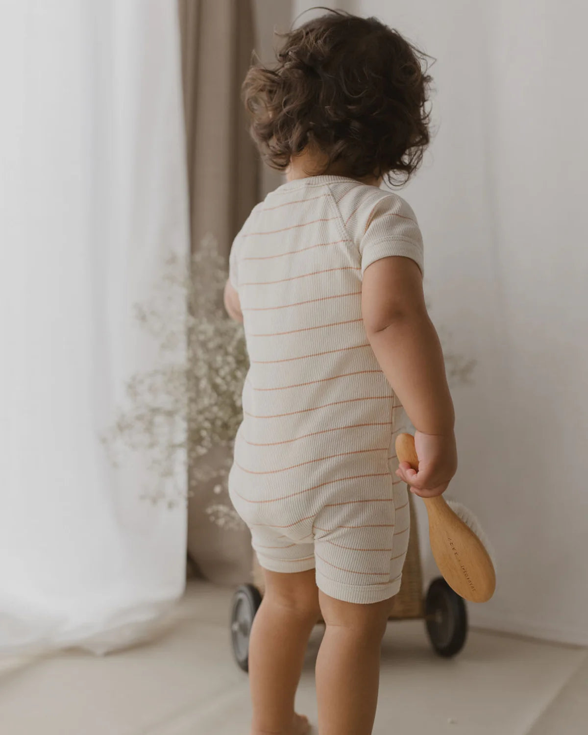 A toddler with curly hair stands facing away, holding a wooden hairbrush. The child is dressed in a light-colored Zip Suit Coco Stripe from SUSUKOSHI, made from organic cotton, and is positioned next to a small wooden wagon. The scene is softly lit with semi-transparent white curtains and adorned with delicate white flowers in the background.