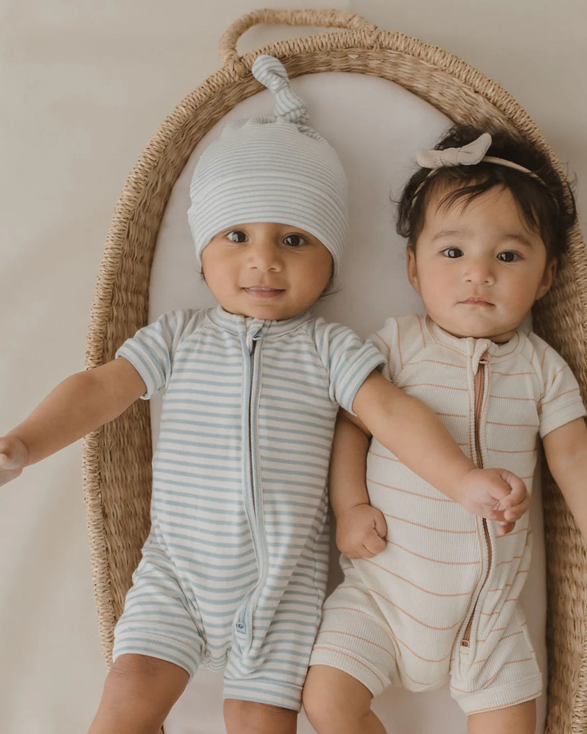 Two babies lie in a wicker basket. The baby on the left wears a light blue and white striped Zip Suit Seaside from SUSUKOSHI, made from organic cotton, complete with a matching knotted hat. The baby on the right sports a beige and white striped onesie with a small bow headband. Both look toward the camera.