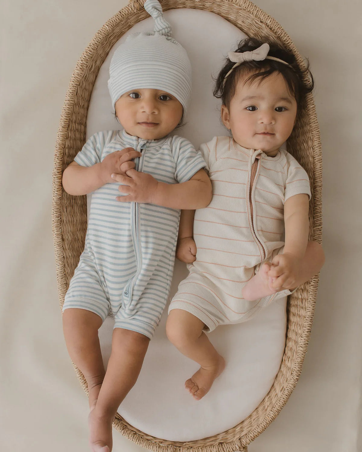 Two babies are lying in a large, oval-shaped wicker basket. The baby on the left is wearing a blue striped Zip Suit Seaside made of organic cotton from SUSUKOSHI, complete with a matching hat. Meanwhile, the baby on the right is dressed in a cream striped onesie and adorned with a bow headband. Both babies have calm expressions as they look up.