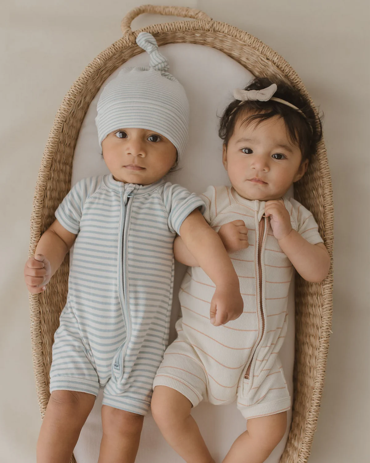 Two babies lie side-by-side in a woven basket. The baby on the left wears the Zip Suit Seaside by SUSUKOSHI, a blue and white striped outfit made of organic cotton, while the baby on the right is dressed in a cream romper with beige stripes and a small bow headband. Both are looking up.