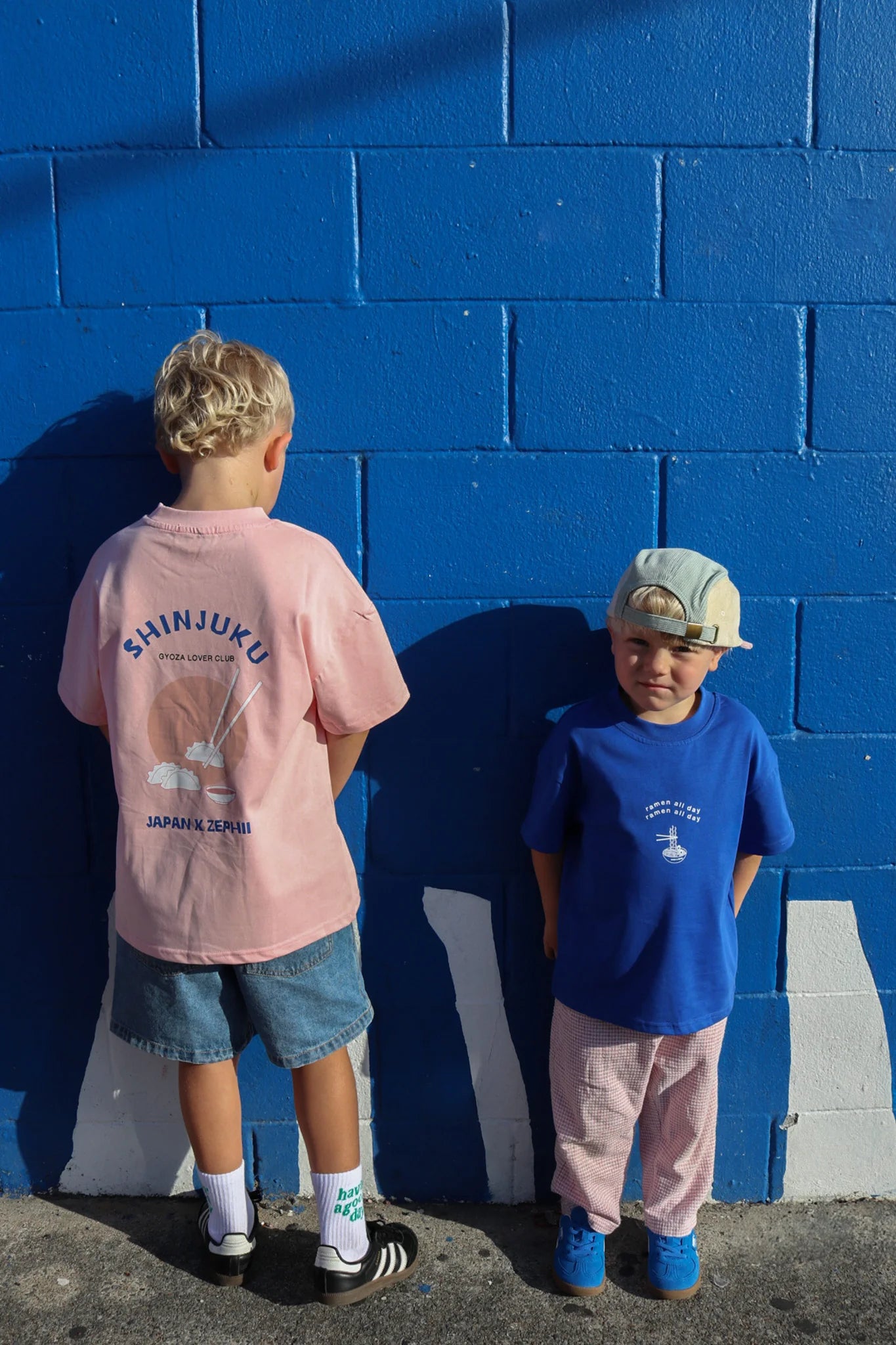 Two young boys express lively Japanese culture against a blue brick wall. The left one in pink shirt and denim shorts faces the wall, while the right one sports an oversized ZEPHII Shinjuku Tee, pink pants, and a backwards cap. Shadows stretch across the ground.