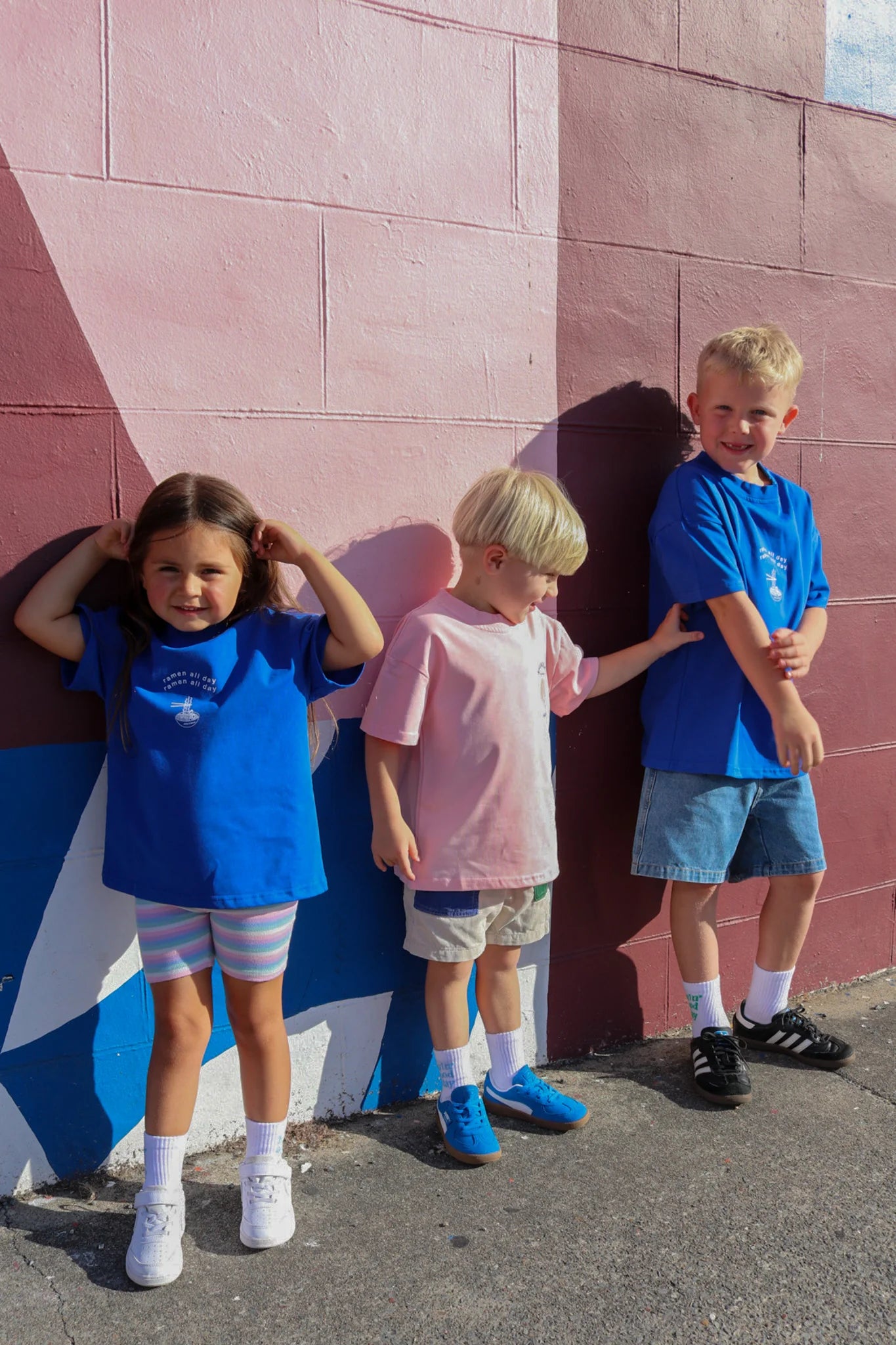 Three children stand by a pink and blue wall. The girl on the left wears a blue shirt and striped shorts with vintage cool; the middle child rocks a ZEPHII Ramen All Day Tee with pink flair, while the boy on the right smiles brightly in a blue shirt and denim shorts.