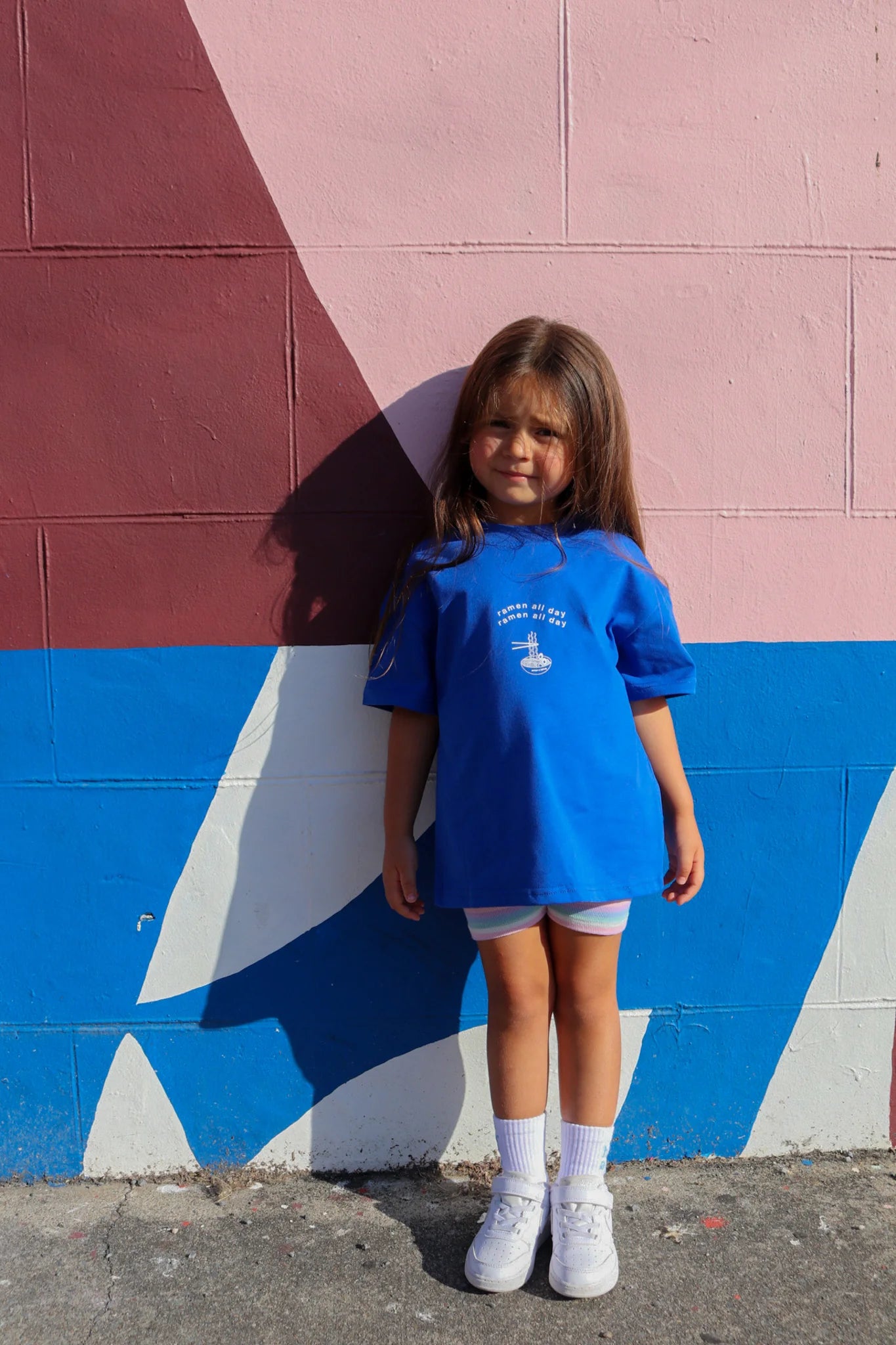 A young girl with long brown hair poses in front of a vibrant mural filled with pink, blue, and maroon geometric shapes. She is wearing a Ramen All Day tee from ZEPHII, paired with light shorts, white socks, and shoes.