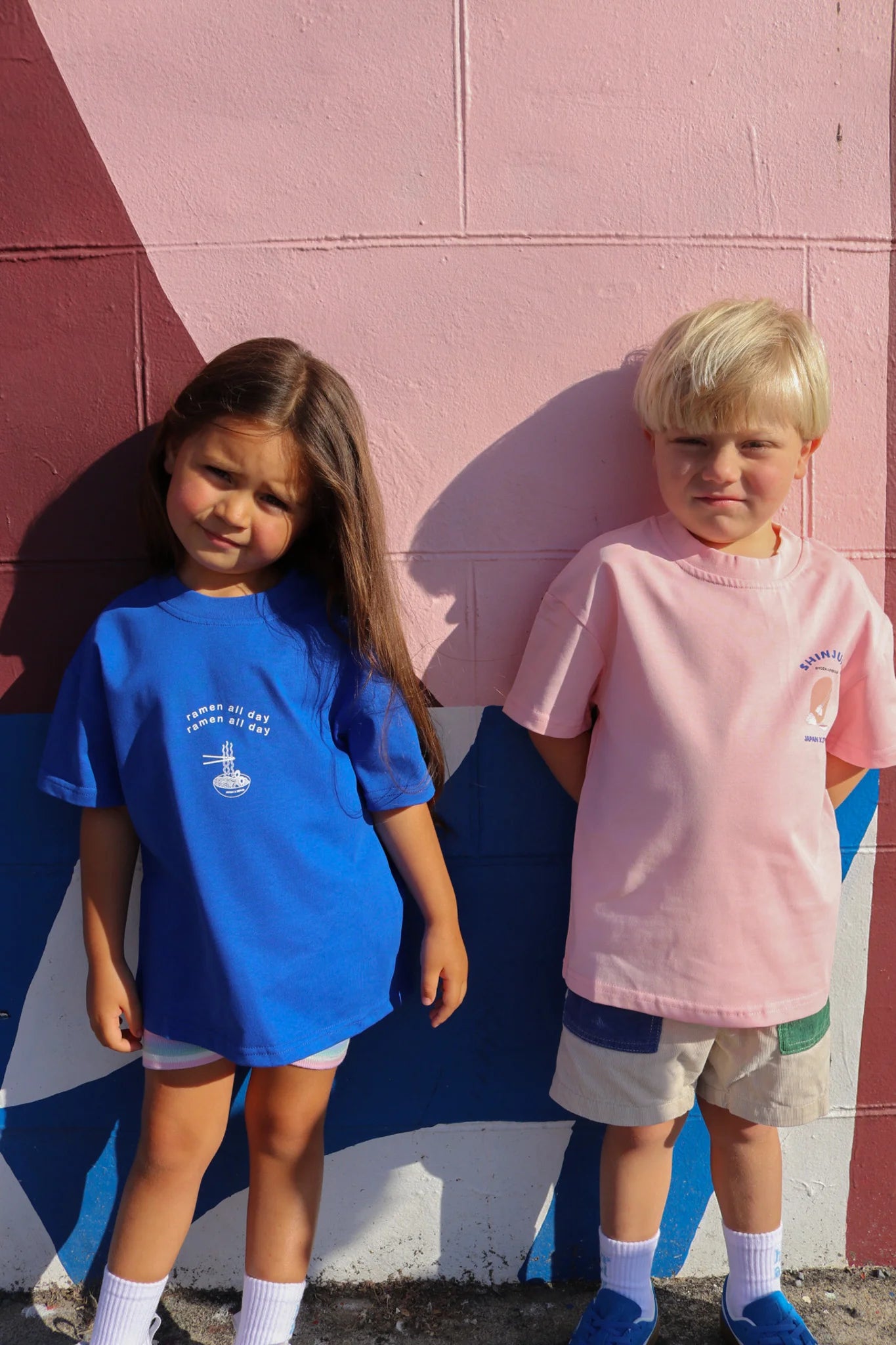 Two kids stand before a vibrant wall. The left child wears an oversized blue tee and shorts, while the right is dressed in ZEPHII's pink Shinjuku Tee and shorts. The wall features geometric patterns in pink, blue, and burgundy, echoing lively Japanese cultural themes.