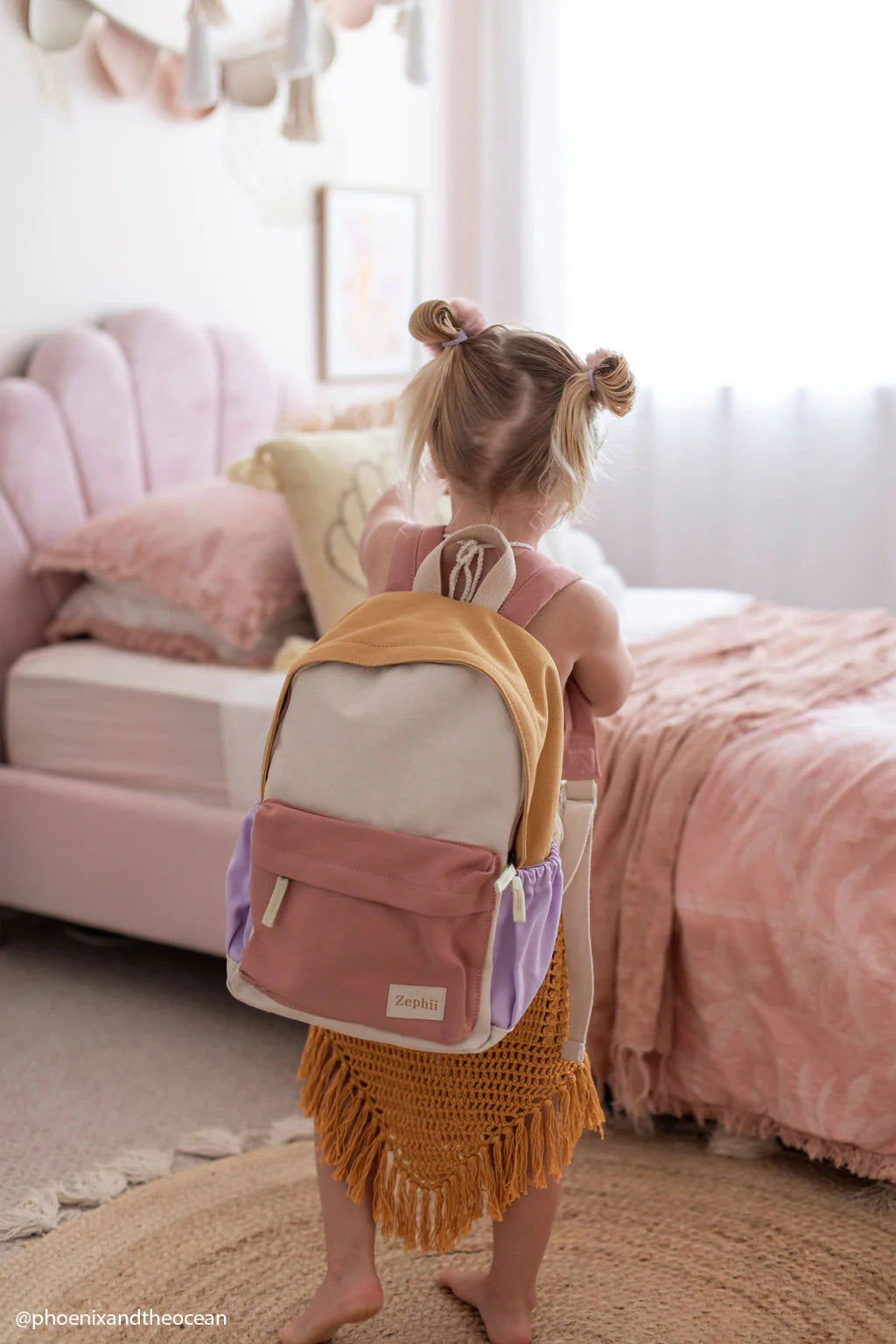 In a warm, pastel-themed bedroom with a pink bed and soft lighting filtering through sheer curtains, a young child with pigtails is dressed in a mustard-colored knitted dress. She's carrying the Mimi Backpack from ZEPHII, known for its versatile design in bold colorways of pink and beige.