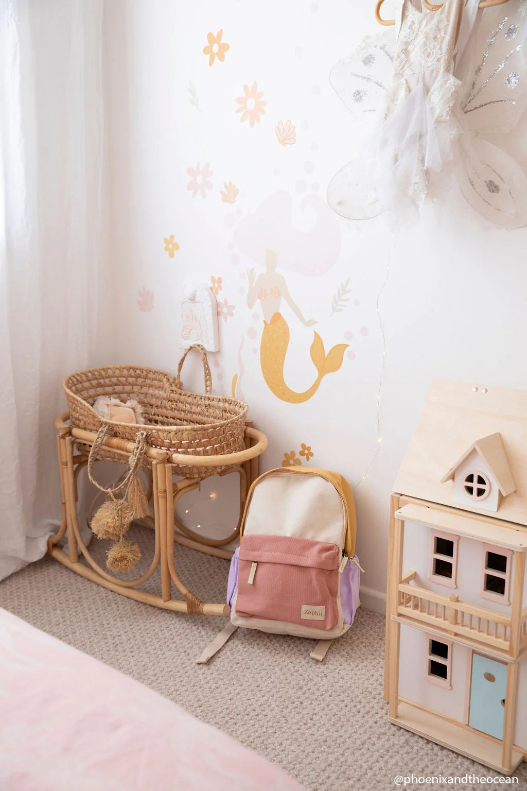 A cozy nursery corner features a wicker bassinet, the versatile Mimi Backpack by ZEPHII in pastel colors, and a small dollhouse on the floor. The wall is adorned with a whimsical mermaid mural and soft, draped curtains, while a hanging robe and string lights add a charming touch.