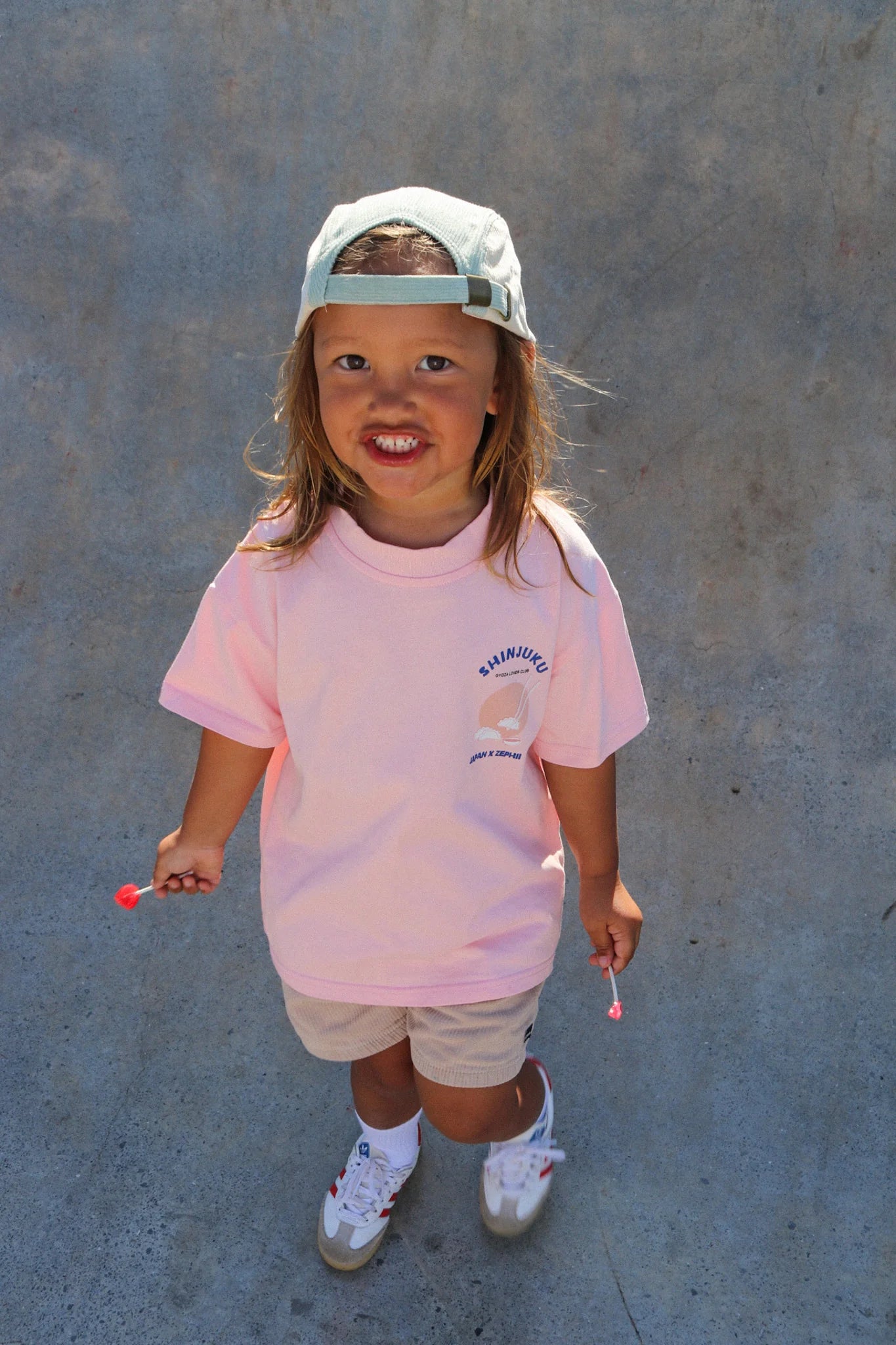 A long-haired child in an oversized vibrant pink Shinjuku Tee by ZEPHII, beige shorts, sneakers, and a backwards cap smiles with two lollipops against a textured grey background.