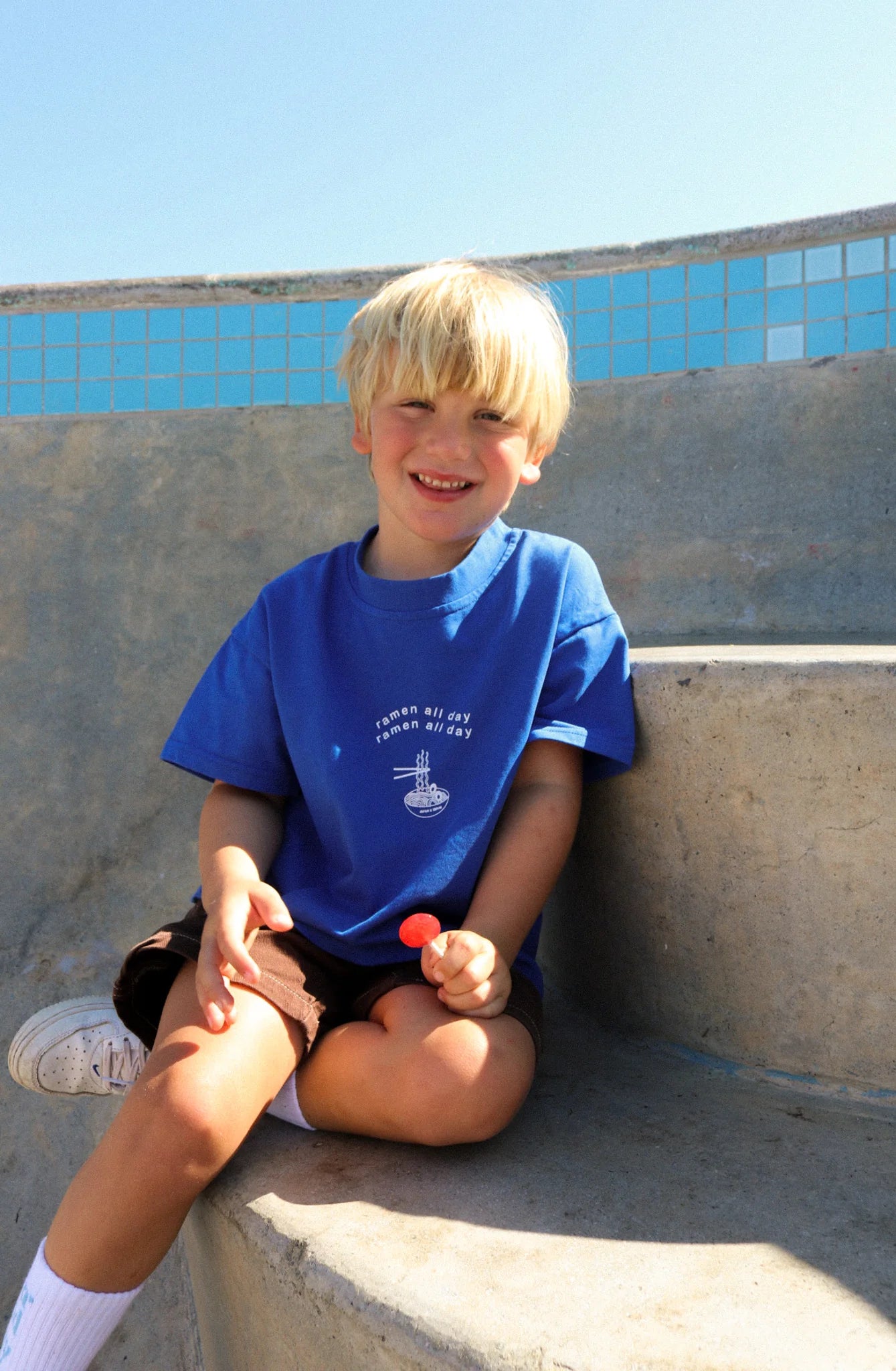 A young boy with blonde hair sits on concrete steps under a clear blue sky, smiling and holding a red lollipop. He's wearing the "Ramen All Day Tee" by ZEPHII, along with brown shorts and white sneakers. The vintage cool scene features a tiled edge in the background.