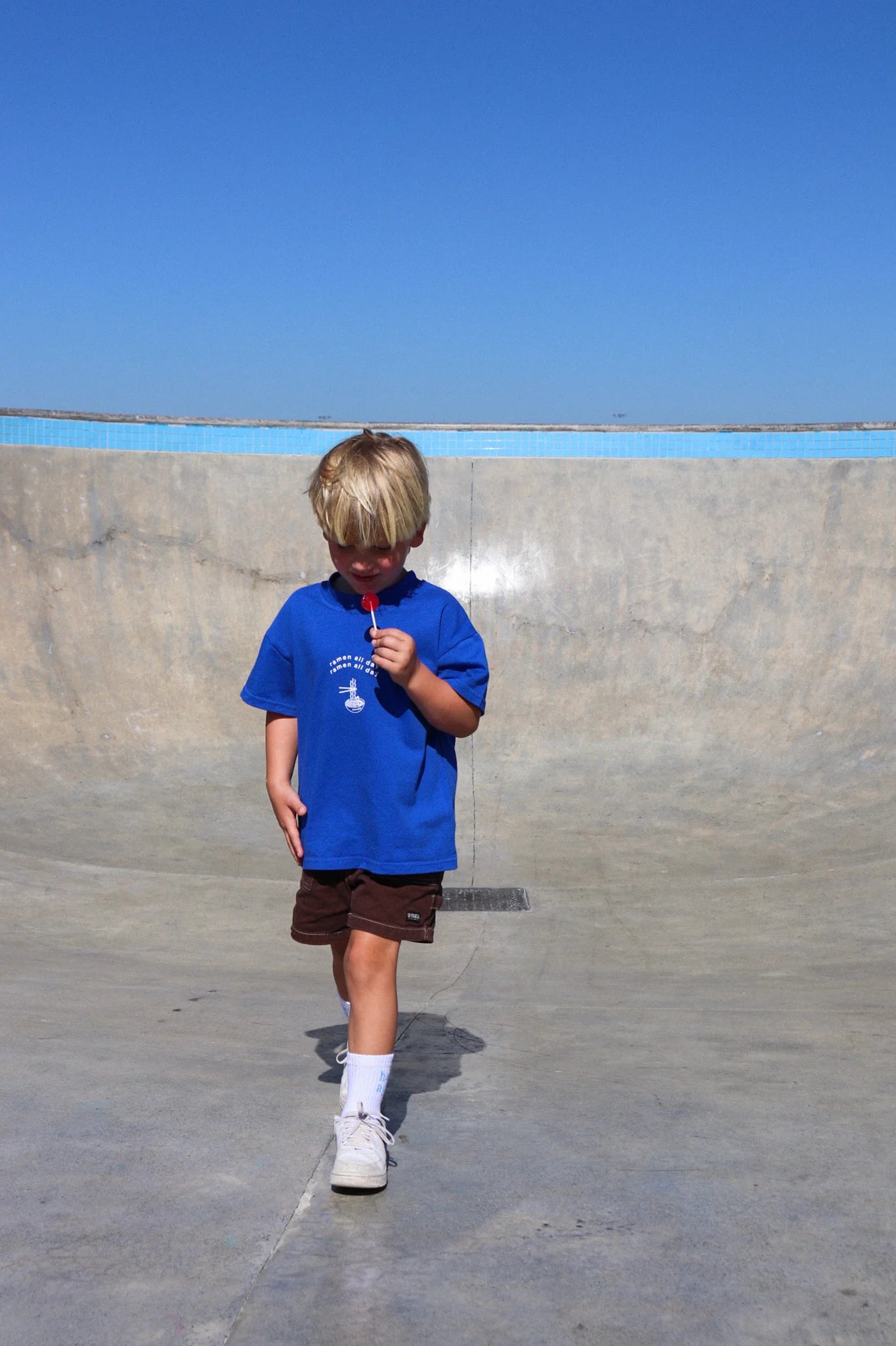 A young blond child strolls through a skate park under a clear blue sky, wearing ZEPHII's Ramen All Day Tee paired with brown shorts. The child, holding a red lollipop and gazing downward, walks past a large concrete ramp.